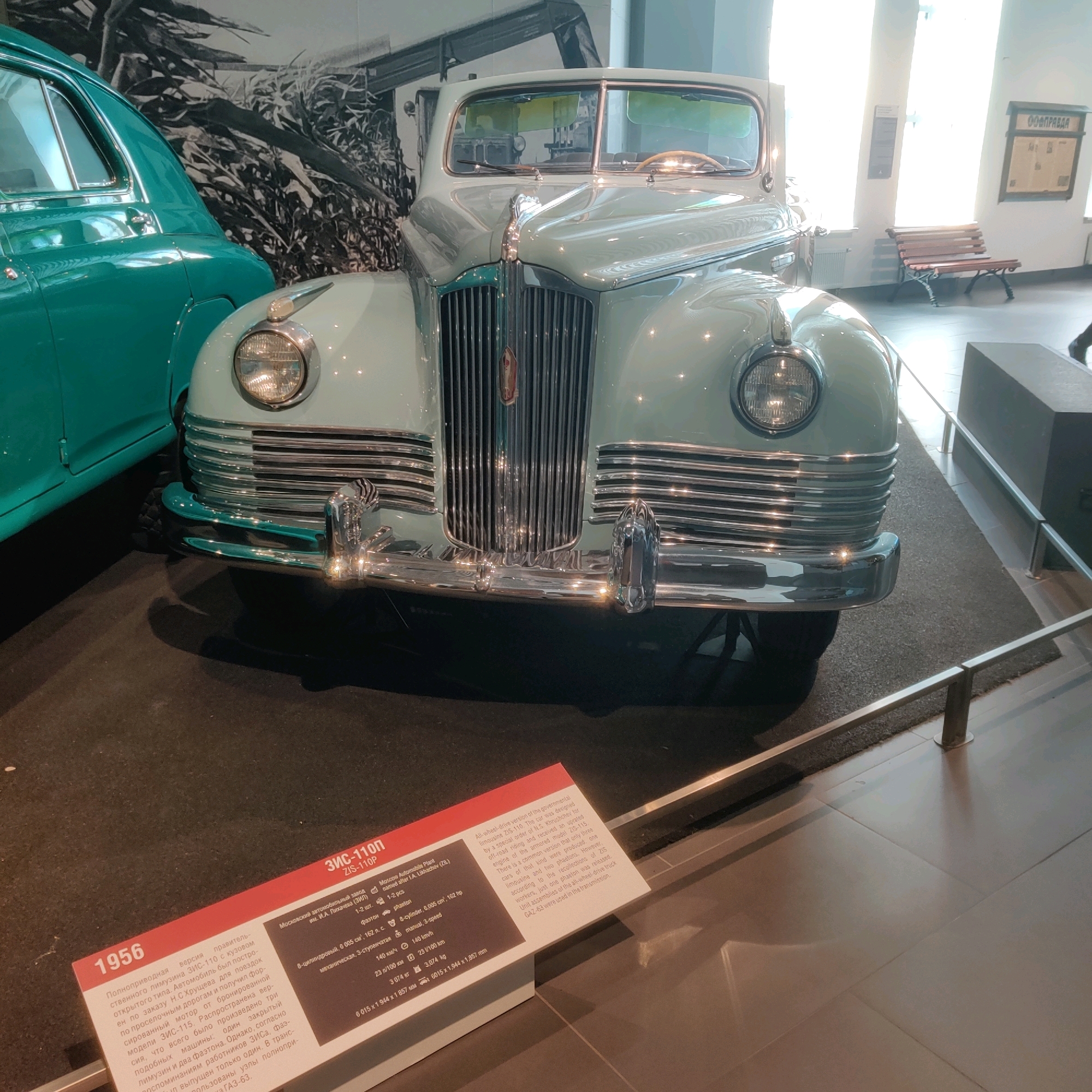 This image shows a light blue 1956 ZIS-110P car on display in the Museum of Military Technology in Verkhnyaya Pyshma, Russia. The car is a governmental limousine with an upgraded off-road engine and an armored version of the ZIS-115. The museum environment is visible, including a dark carpeted floor, polished tiled floor in the background, and exhibition barriers. A second, teal-colored car is partially visible to the left of the ZIS-110P. A display sign with text in Russian and English is positioned in the foreground, providing information about the car. In the background, a wooden bench and a framed display are visible on a wall. The lighting suggests an indoor exhibition space with natural light coming from windows.
