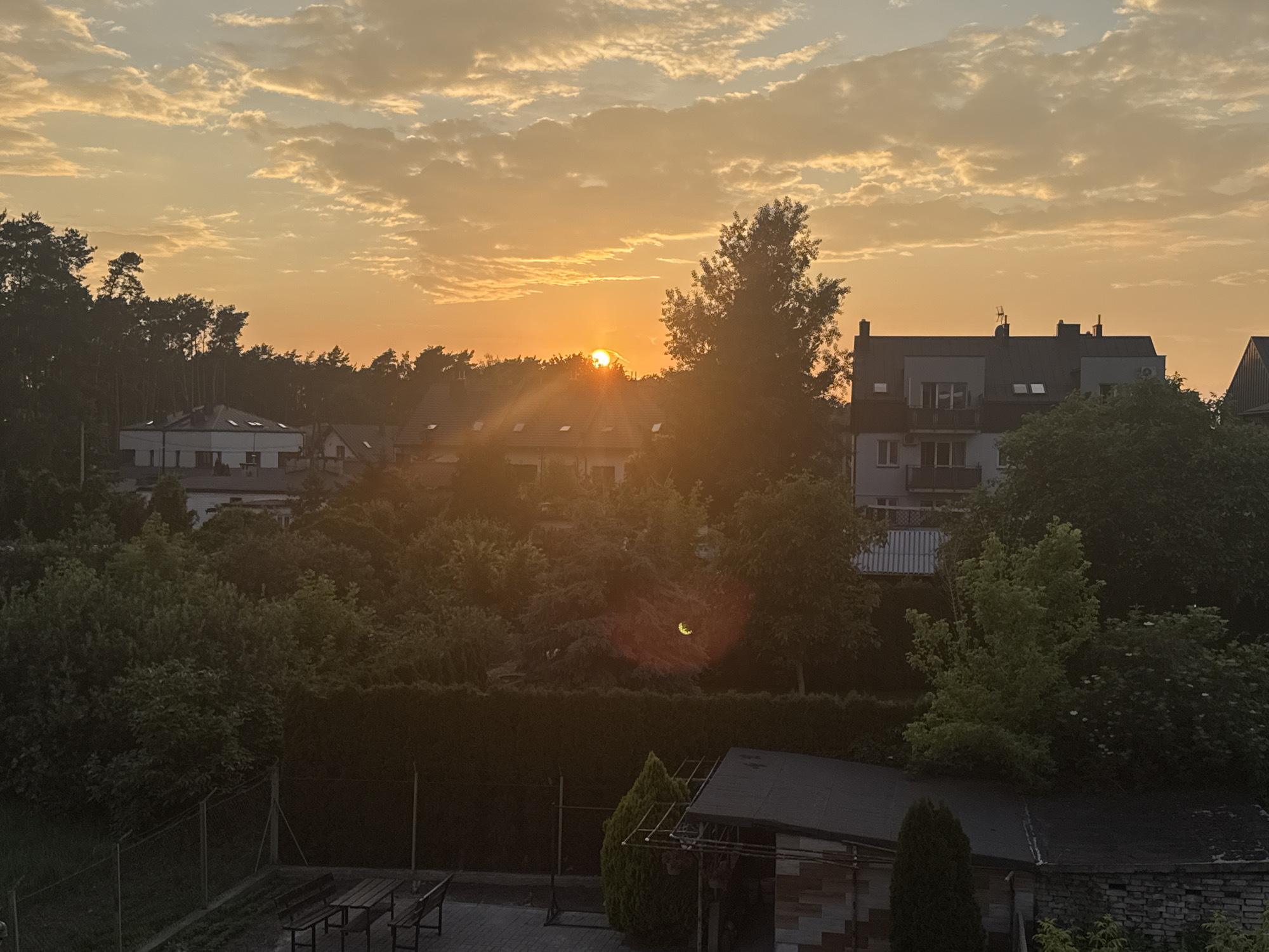The image depicts a serene residential area in Marki, Poland, during what appears to be a sunrise or sunset. The sky is filled with soft, elongated clouds illuminated by the warm, orange glow of the sun, which is partially obscured by trees. In the background, several multi-story buildings with pitched roofs are visible, some featuring balconies and dormer windows. The foreground shows a fenced yard with a picnic table and benches, surrounded by dense green foliage and trees. A small, simple shed with a dark roof and brick base is also present. The scene is characterized by a peaceful, suburban atmosphere, with no people or animals visible.