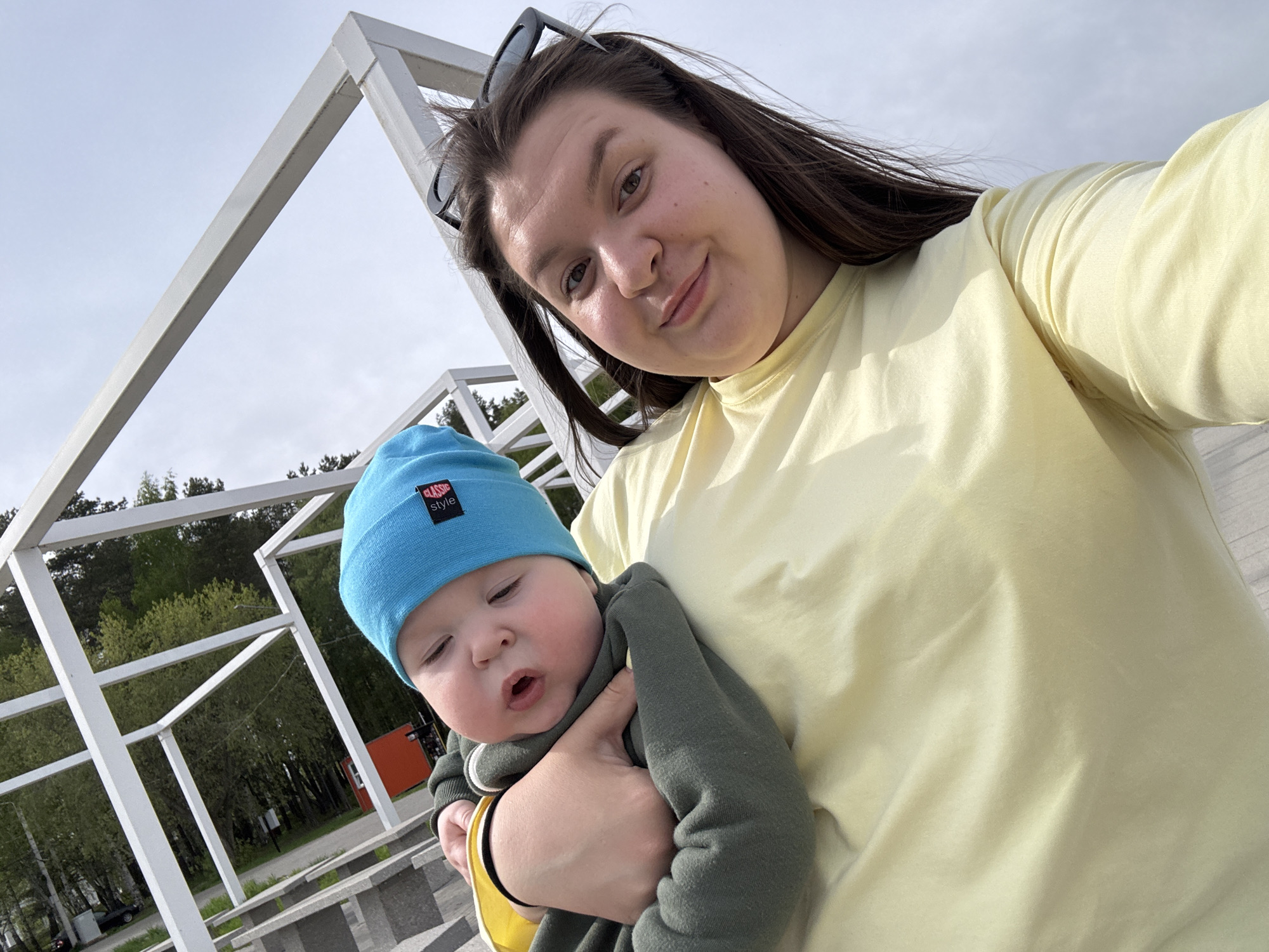 A woman with brown hair and sunglasses atop her head is holding a baby wearing a blue hat with the words "CLASSIC style" on it. The woman is wearing a light yellow shirt. The background shows a white metal structure, trees, and a cloudy sky. The picture was taken in Chaikovsky, Russia.