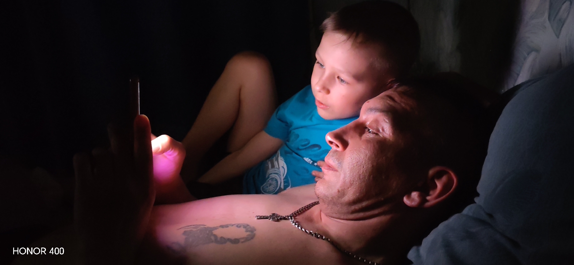 In a dimly lit indoor environment, a man and a young boy are seen looking at a mobile phone held by the man. The man is lying down, shirtless, with a silver chain necklace and a tattoo on his chest. The boy is wearing a blue t-shirt and is seated behind the man. The light from the phone screen illuminates their faces. The "HONOR 400" watermark is visible at the bottom left of the image. The location is in the city of Noviy, Russia.