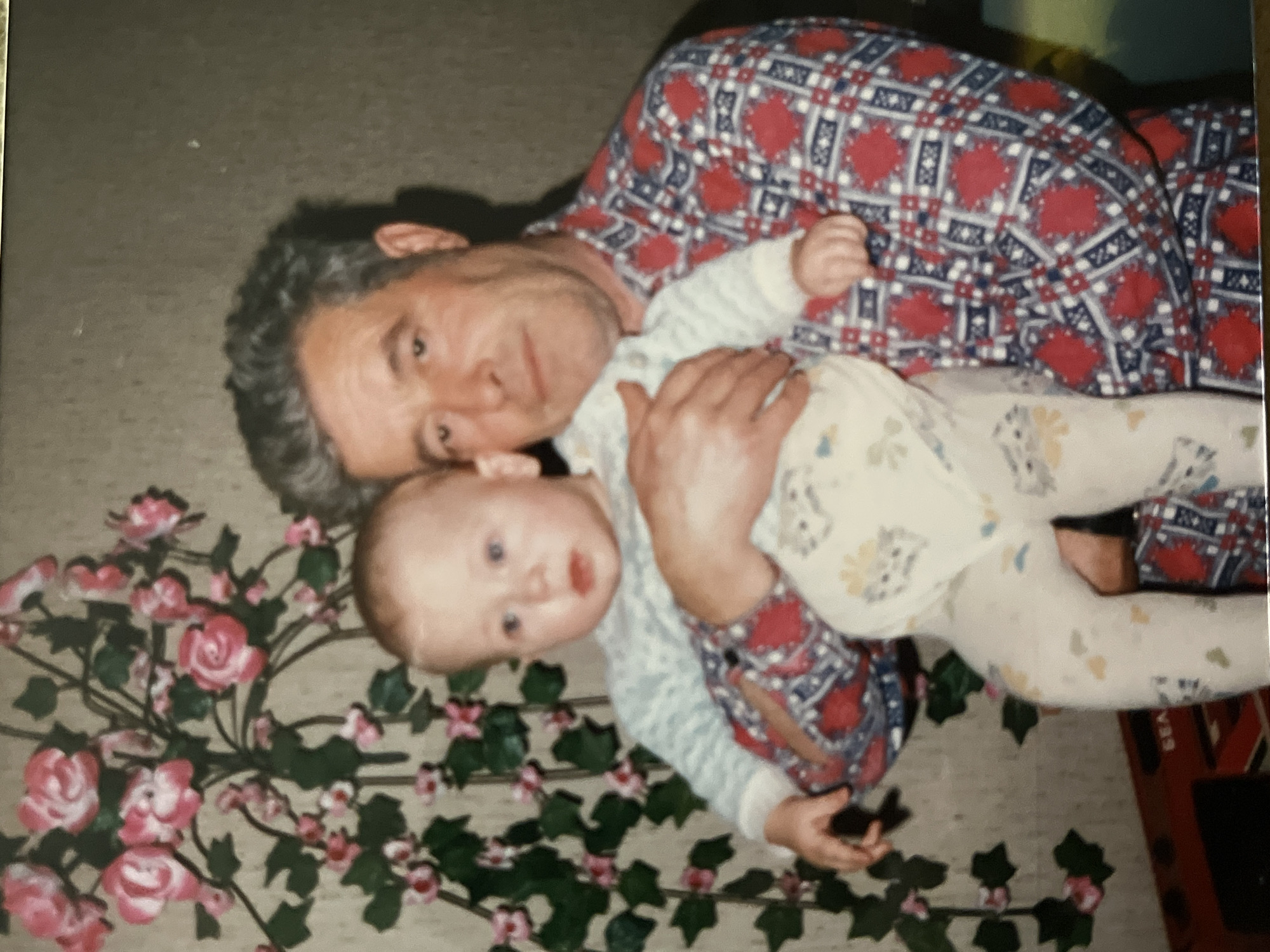 This indoor photograph, taken in Minsk, Belarus, features an adult man holding a baby. The image is oriented horizontally, depicting the two subjects lying on a light-colored surface.
The adult, appearing to be a man, has graying, curly hair and a gentle, slightly smiling expression. He is wearing a long-sleeved shirt with a distinctive pattern of red, blue, and white geometric shapes and plaid elements. He is cradling a baby, who has light hair, blue eyes, and a slightly open mouth, looking towards the viewer with a curious expression. The baby is dressed in a light-colored onesie adorned with subtle, faded patterns that resemble animals or small objects.
Around the subjects, primarily on the left and lower portions of the frame, an artificial floral garland is arranged on the surface. This garland features clusters of pink roses and green leaves. A dark shadow is visible behind the adult's head. On the far right edge of the image, a glimpse of a red and black object is present, with faint vertical text that appears to include the letters "SEA". The overall scene conveys a tender and intimate moment between the adult and the baby.