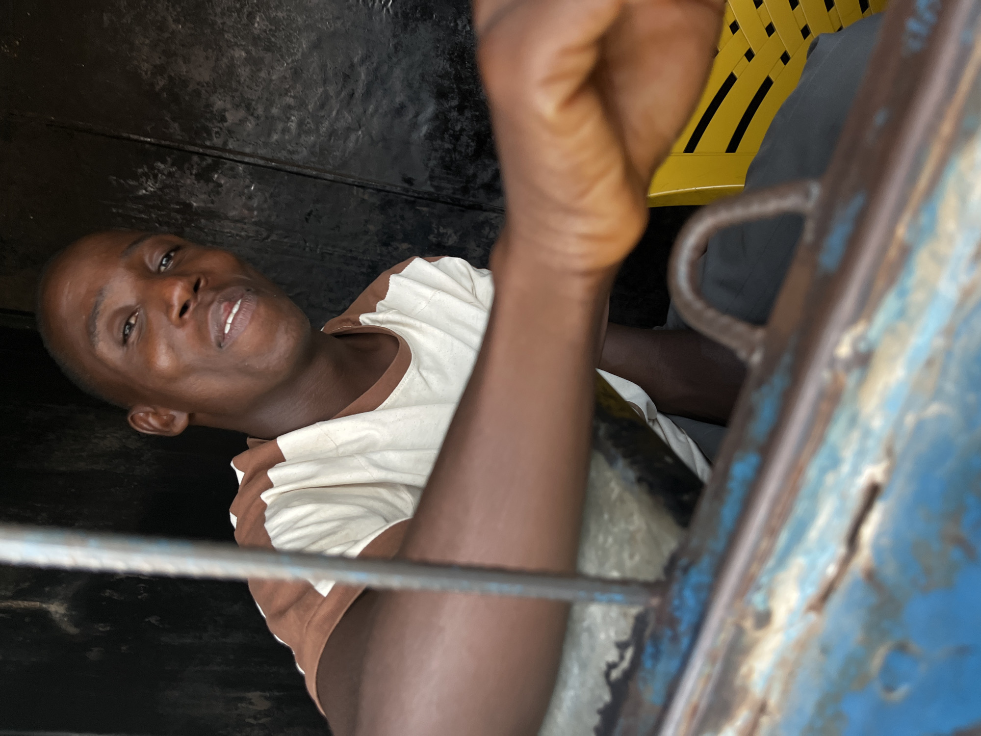 A man with dark skin and short-cropped hair is looking through a blue-painted metal barred window. He is wearing a white t-shirt with brown sleeves and collar. His right arm is raised and partially obscuring the view. Behind the man is a yellow woven plastic chair and a person wearing grey pants. The location appears to be indoors. There are no other discernible details to confirm the specific location within the city of Jos, Nigeria.