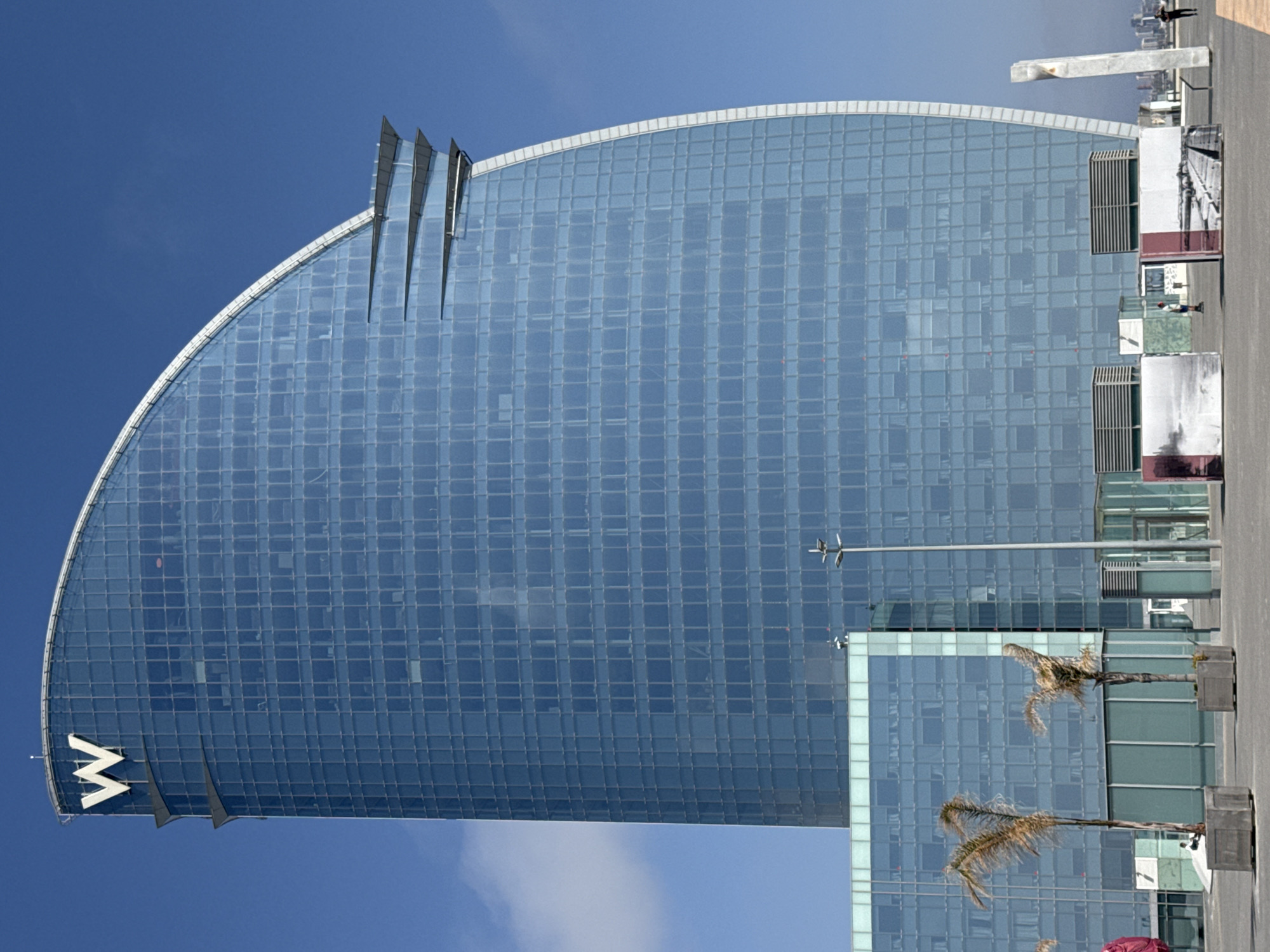 The image shows the W Barcelona Hotel, located in Barcelona, Spain. The hotel is a modern glass structure shaped like a sail. The building is largely blue with numerous windows. In the bottom left, there is a large "W" logo. The sky is blue, and the surrounding area includes palm trees and other structures.