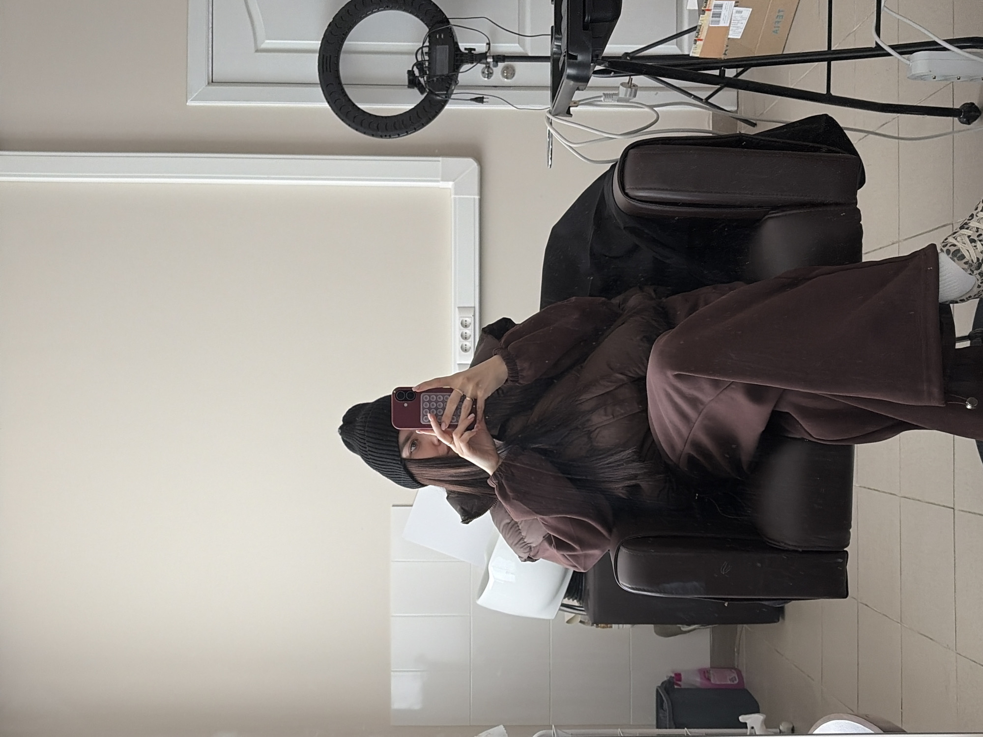 An indoor, mirror-reflection image depicts a young person seated in a dark brown, padded salon-style chair, taking a selfie with a smartphone. The person is wearing a dark beanie, a dark brown puffer jacket, and dark brown wide-leg pants. Their long dark hair partially covers their face as they hold a dark smartphone with visible camera lenses towards the reflective surface. One foot, wearing a light-colored shoe with a distinct leopard print, is visible.
The setting appears to be a studio or salon in Chaikovsky, Russia. To the left of the person is a large black LED ring light mounted on a stand, with various wires and cables connected and visible. Behind the ring light, a white power strip or multiple electrical outlets are affixed to the light beige wall. Further back and slightly above the person, a white panel door is seen. A white roller blind covers a window behind the chair.
Numerous black and white cables are strewn around the ring light stand. A cardboard box, partially visible on a shelf behind the stand, bears the mirrored text "AIRT". On the tiled floor to the right of the chair, several items including bottles (one pink) and a black bag or container are visible. The overall impression is a casual moment captured in a functional indoor environment, possibly during a waiting period or break. The artificial lighting from the ring light suggests it could be any time of day.