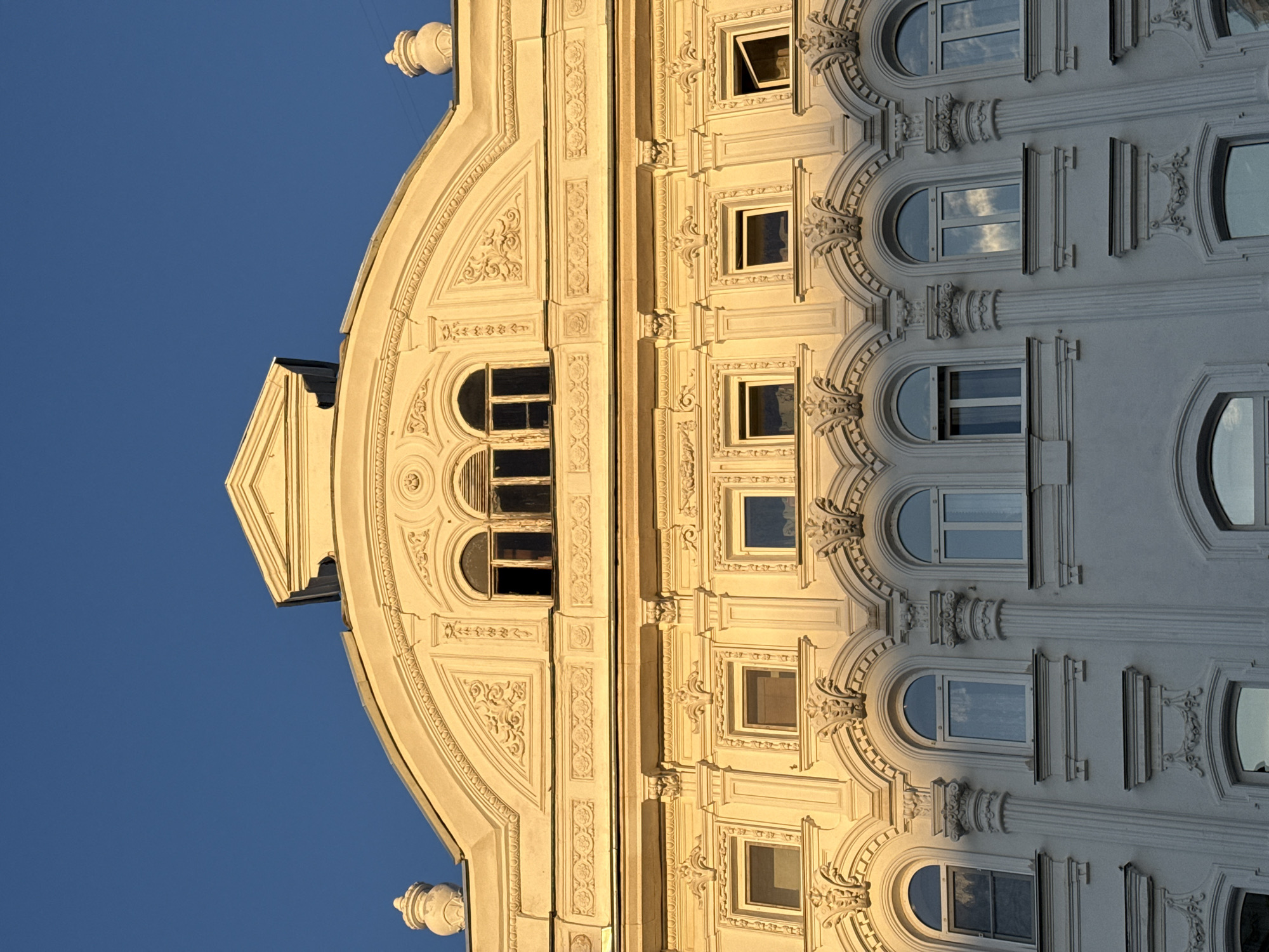 This is a close-up, low-angle, eye-level shot of the facade of a white, ornate building in Nizhny Novgorod, Russia, illuminated by the warm, golden light of the setting sun. The building features multiple stories with numerous windows of various sizes and shapes. The upper stories display elaborate decorative elements, including friezes, pediments, and sculpted ornamentation around the windows. Some windows are open, while others are closed, and reflections of clouds are visible in some of the glass panes. The clear blue sky serves as the background. The image lacks any human presence or activity.