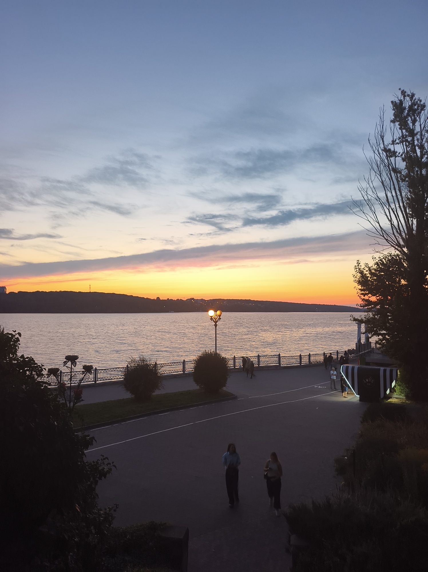 The image depicts a twilight scene along the Ternopil Pond, also known as Ternopil Lake, in Ternopil, Ukraine. The sky exhibits a gradient of colors, transitioning from a pale blue at the top to vibrant orange and yellow hues near the horizon, indicating sunset or sunrise. A distant, silhouetted treeline forms the backdrop across the water. Along the water's edge, a promenade is visible, featuring a decorative railing, lampposts, and sparse vegetation. Several individuals are scattered along the promenade, some walking, others standing. In the foreground, two young women are walking on a paved pathway. The overall atmosphere is serene, with the water appearing calm and reflecting the colors of the sky. The scene captures a public space in Ternopil during a transitionary period of light.