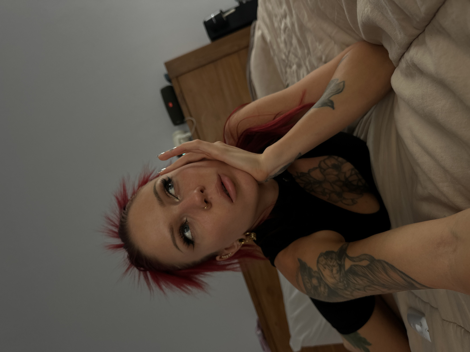A fair-skinned person with red, spiky hair is lying on a bed covered with a light beige blanket. The person is wearing a black top and has multiple tattoos on their arms and chest. The person's left hand is touching their face. They have piercings in their nose and ears. In the background is a wooden dresser and a light gray wall. The location is not specified and cannot be inferred from the image.