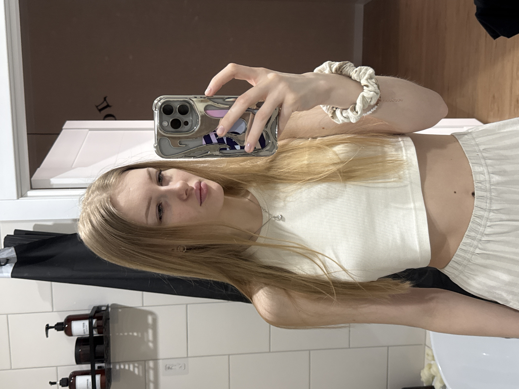 A blonde woman is taking a selfie in a bathroom with white tiled walls. She is wearing a white tank top and striped pants. She is holding a phone with a decorative case. Bathroom amenities such as a soap and lotion dispenser, and a black garment are visible. There is no indication of the location being Perm, Russia.