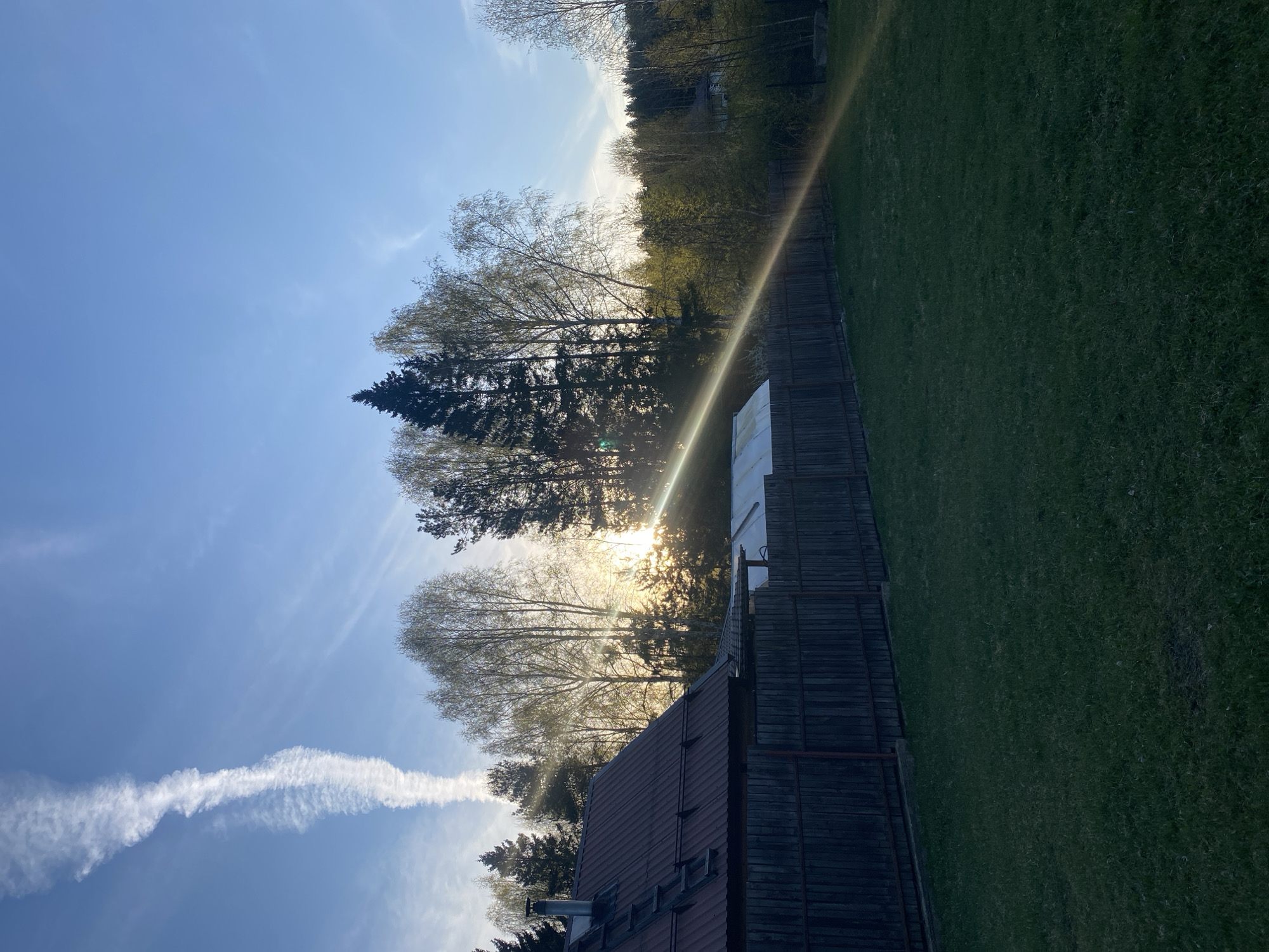 The image shows a scene in Verkh-Neyvinsky, Russia, at dusk. The sky is light blue, with the fading contrail of a jet airplane visible in the lower left. The sun is setting or rising behind a stand of trees. A dark green lawn is on the right side of the image and is bordered by a wooden fence. A building with a red tiled roof is at the bottom of the image. Sunlight is refracted through the trees.
