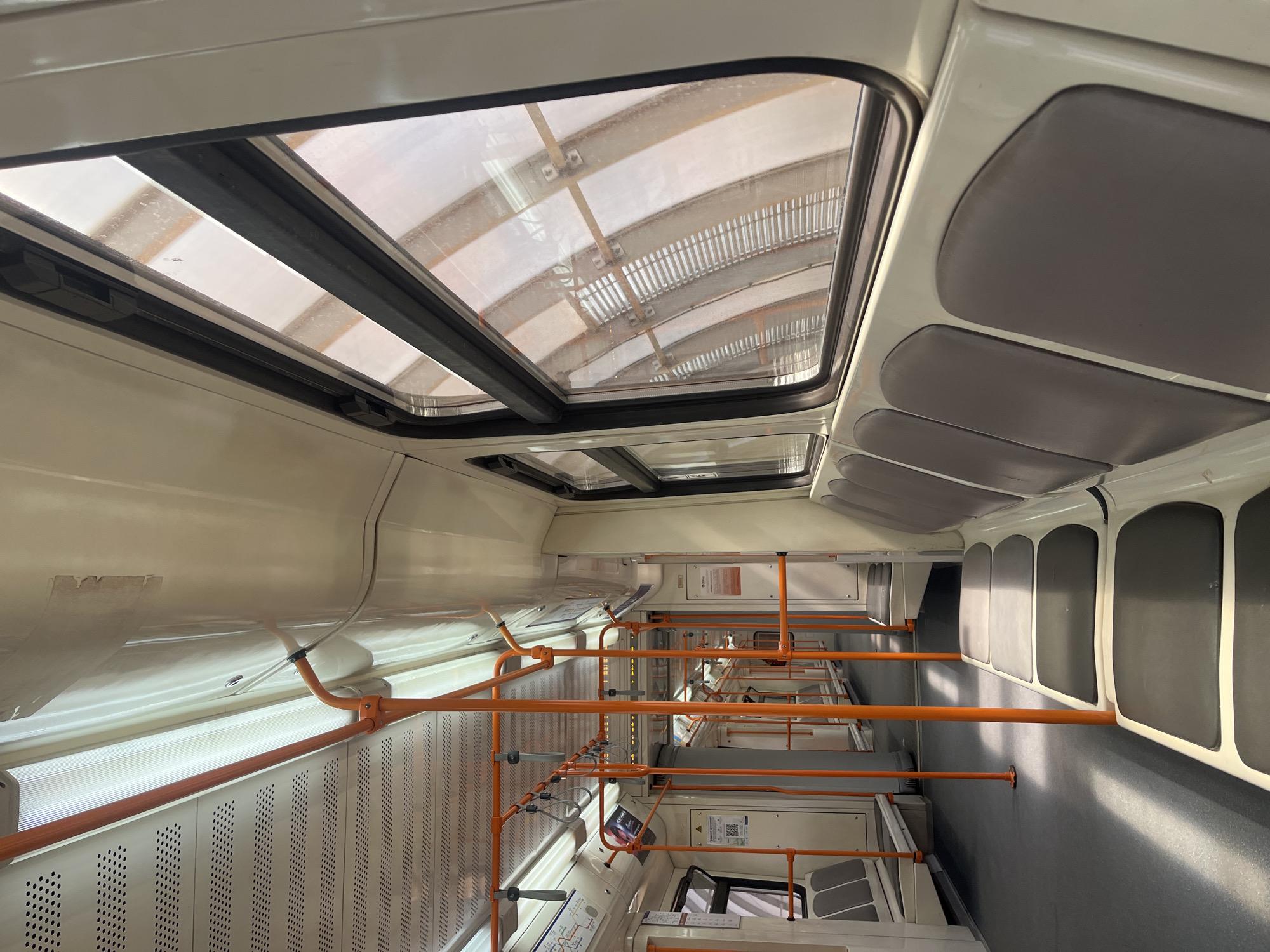 The image shows the interior of a light rail vehicle, specifically a tram, in Sofia, Bulgaria. The tram features a white and gray interior with orange handrails and overhead windows. The roof has rectangular skylights. Gray padded seats are along the right wall of the vehicle. The tram appears empty and stationary. The environment includes the city infrastructure seen through the windows, suggesting an urban setting.
