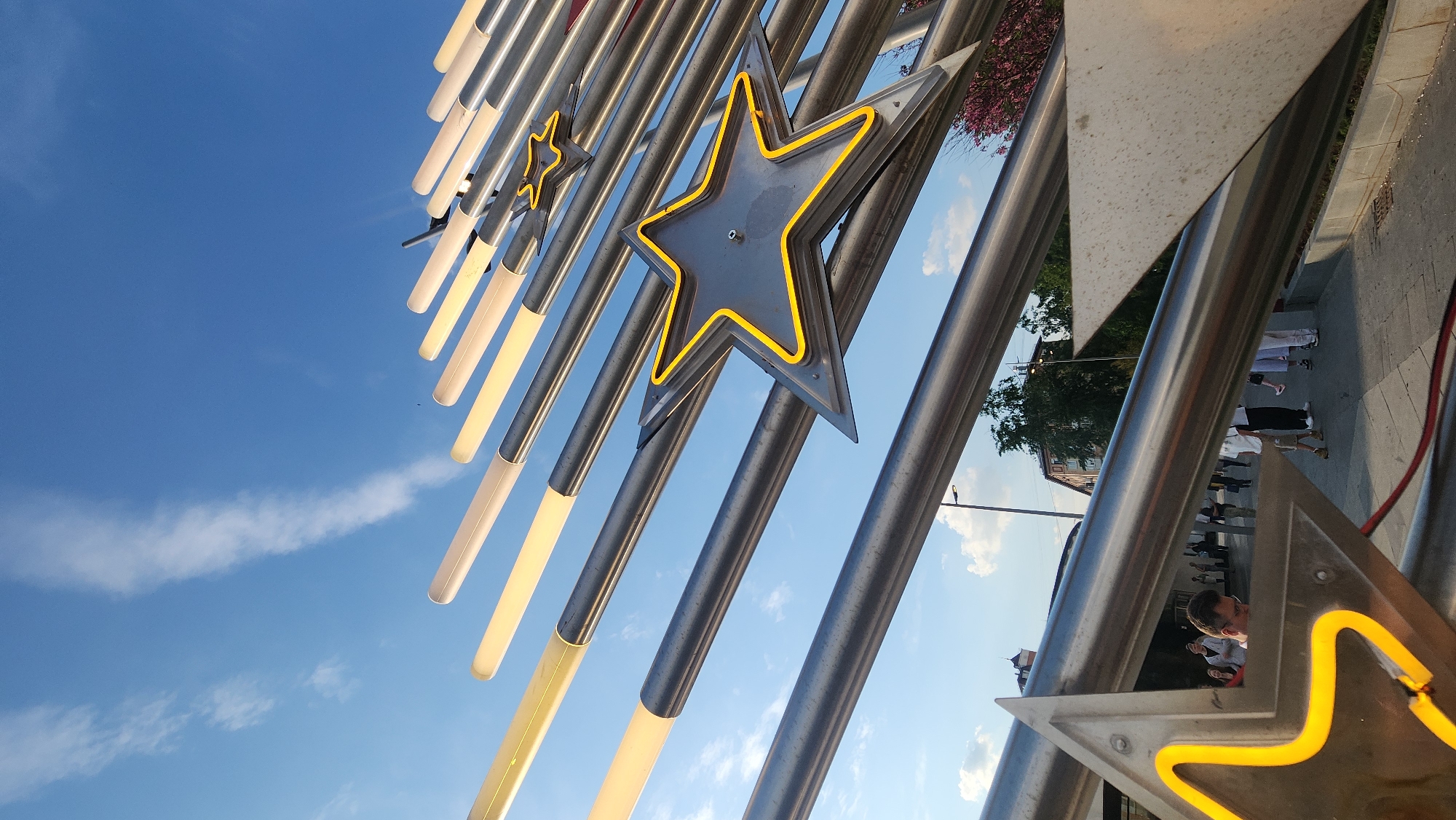 The image depicts a public art installation in Moscow, Russia. The installation features metal tubes arranged diagonally, some of which are illuminated with a warm yellow light. Several large metal star shapes, outlined with yellow neon, are mounted on the tubes. The background is a clear blue sky with scattered clouds. On the ground, pedestrians are walking along a sidewalk, some wearing light-colored clothing. Buildings and trees can be seen in the distance.