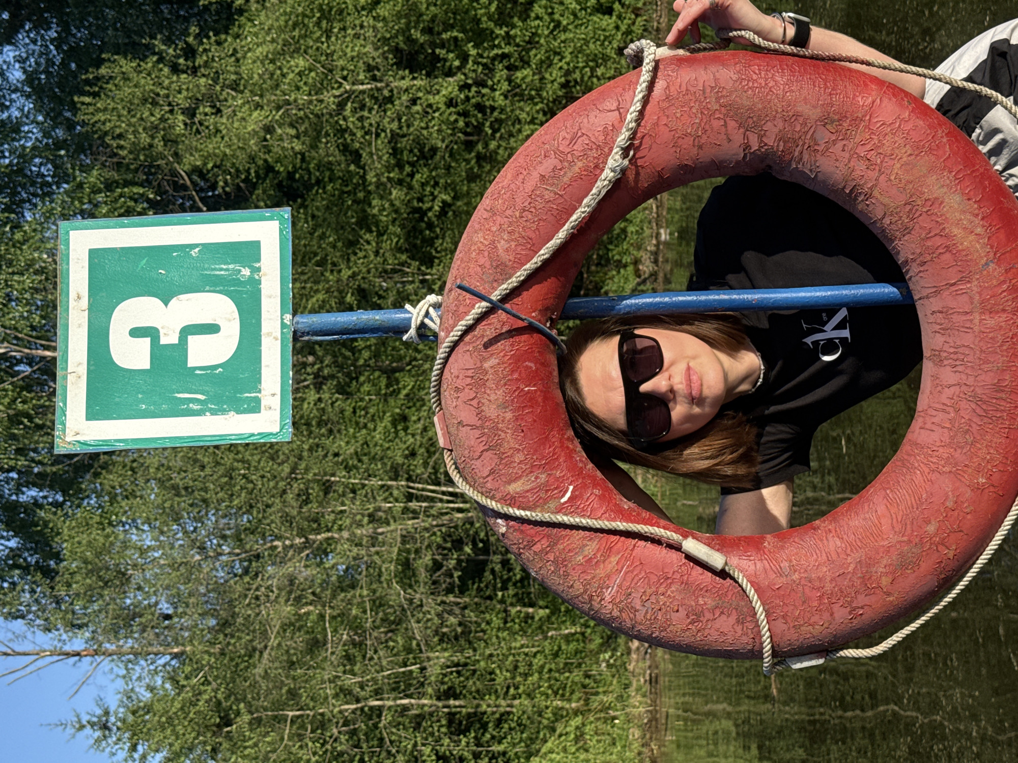 A medium shot shows a woman wearing sunglasses and a black T-shirt with white lettering, partially obscured by a circular red life preserver with a light-colored rope attached. The life preserver is mounted on a blue pole. Adjacent to the life preserver is a square green sign with a white border and the number "3" in white. The woman is looking towards the camera. Behind her, blurred trees and water are visible. The location is in Chaikovsky, Russia.