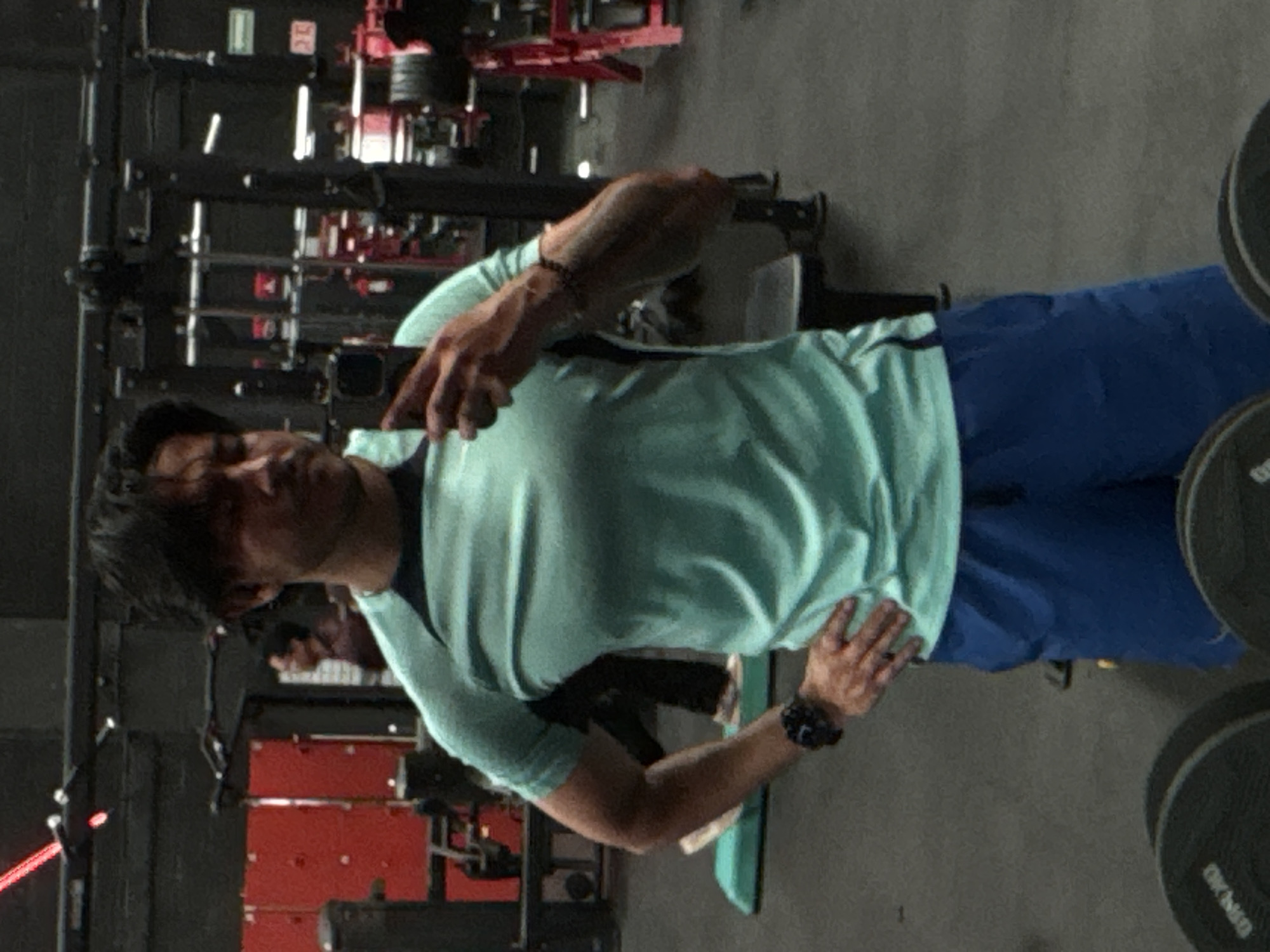 A person in a light green t-shirt and blue shorts is taking a selfie in a gym. The person has dark hair and is wearing a bracelet on one wrist and a watch on the other. Gym equipment is visible in the background. The location is not identifiable as the city of Puebla, Mexico, from the provided image.