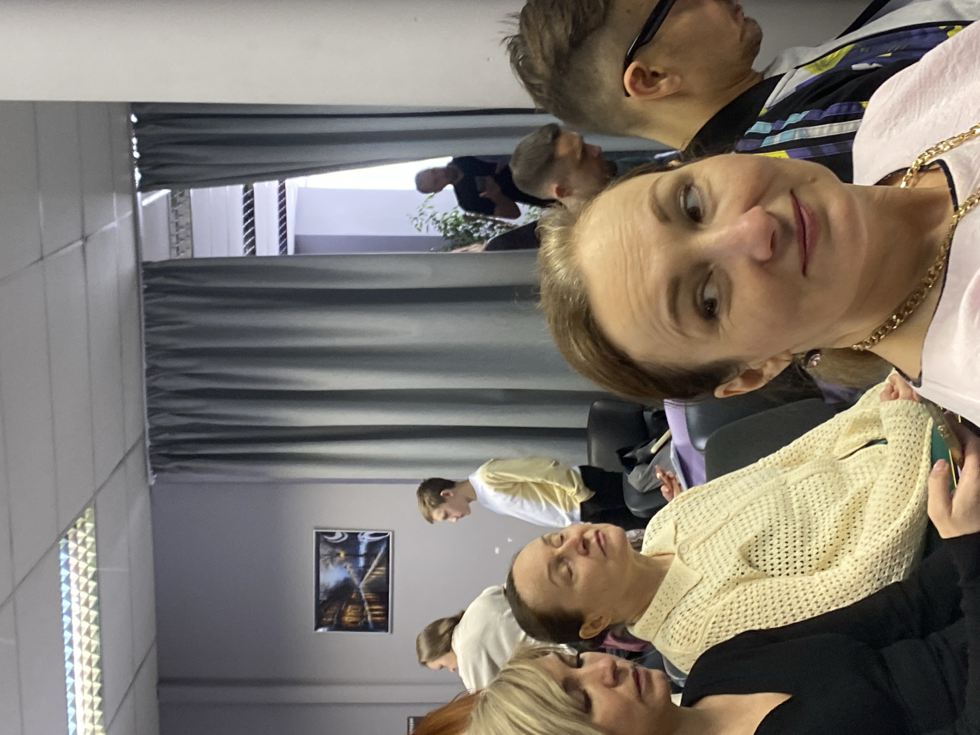 Here is a detailed description of the image:
**People:** The image depicts a group of individuals in a room. The main focus is on a woman in the foreground, wearing a pink shirt and a gold chain. She is looking towards the camera. Behind her, there is a man in a patterned shirt and glasses. Other people are seated in the background, with varied clothing.
**Objects:** Various objects are visible in the scene, including chairs, a picture on the wall, and a plant. A cardigan is worn by one of the people behind the primary person.
**Actions and Interactions:** The subjects appear to be sitting and possibly participating in an event or meeting. The woman in the foreground seems to be the focus of attention, likely because she is looking at the camera.
**Environment:** The setting seems to be an indoor space, possibly an office or a meeting room. There are gray curtains on one side of the room. The walls are a light color. The lighting is subdued, suggesting a comfortable environment.
**Location:** Unknown, Unknown.