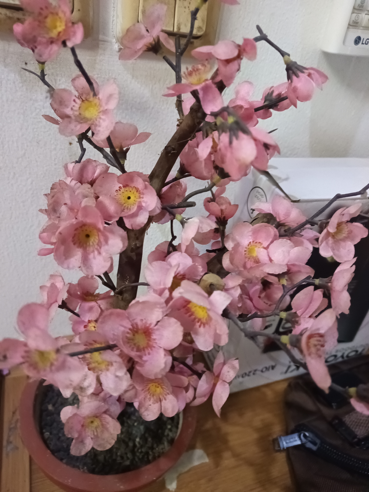 The image shows a close-up view of an artificial pink cherry blossom tree in a reddish-brown pot. The tree has several branches adorned with numerous pink flowers, each with a yellow center and a dark pistil. The background is a textured white wall. To the right, partially visible, are some white boxes with text and a brown bag with a zipper. The lighting appears to be indoor. This scene is likely located in a residential or commercial indoor space in North Jakarta, Indonesia.