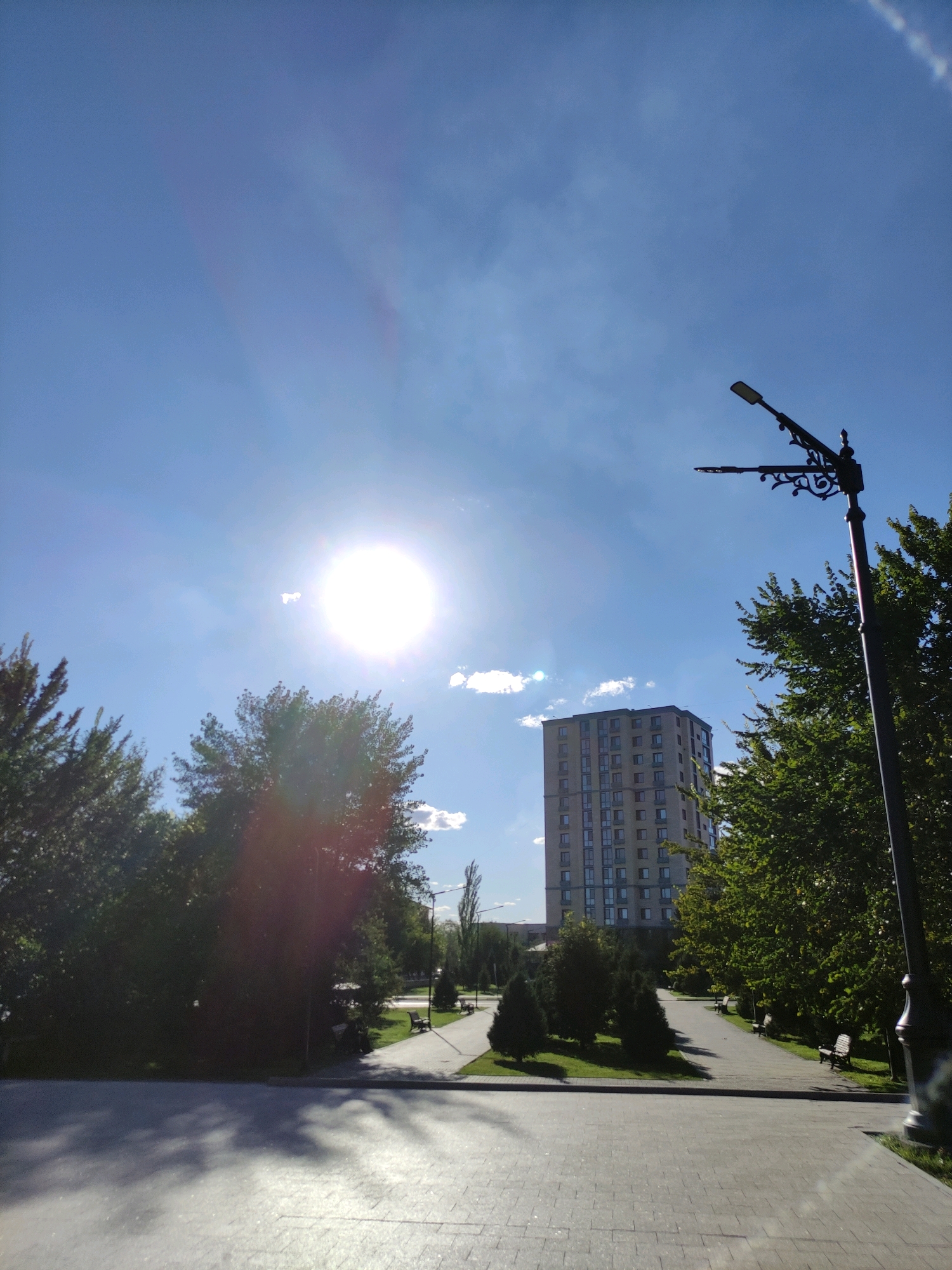 The image depicts a park-like setting with a paved walkway leading towards a tall, multi-story building. Lush green trees line the walkway, casting shadows on the ground. A bright sun is visible in the sky, causing a lens flare. A decorative lamppost stands prominently on the right side of the frame. In the distance, a modern apartment building rises above the trees. Benches are visible along the walkway, suggesting a public space for relaxation. The scene appears to be in the city of Semey, Kazakhstan, indicated by the architectural style of the building and the presence of urban park infrastructure. The environment is outdoors, under a clear blue sky with a few scattered clouds. No people or specific actions are discernable.