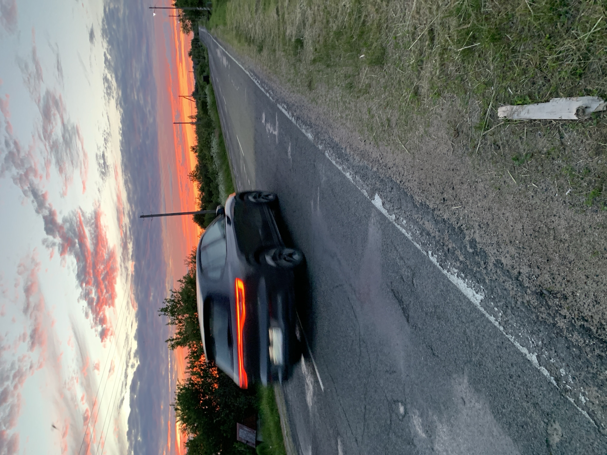 The image captures a rural road scene in Відзаўскі сельскі Савет, Belarus, at sunset or dusk. A dark-colored SUV, possibly black or dark grey, is depicted in motion, traveling away from the camera on the right side of an asphalt road. The vehicle exhibits significant motion blur, particularly on its wheels and lower body, with its rear LED taillights glowing brightly in a horizontal strip design. The license plate is unreadable due to blur.
The sky is a prominent feature, displaying a gradient of colors from light blue and grey at the top to vivid reds, oranges, and pinks at the horizon, indicating a strong sunset. Scattered clouds are illuminated by the setting sun. Power lines are visible against the sky in the upper left quadrant.
The asphalt road, appearing somewhat worn with cracks and patches, stretches into the distance, ascending slightly and curving gently. A faded white solid line marks the right edge of the paved surface. On the left side of the road, dense green vegetation, including trees and bushes, lines the roadside, partially obscuring a small, dark brown wooden structure. Multiple utility poles, primarily wooden, are visible on both sides of the road, extending towards the horizon. The right shoulder consists of a narrow strip of gravel and dirt, transitioning to sparse grass with scattered dry debris, including a short, light-colored wooden plank lying horizontally in the foreground. No individuals are present in the scene.