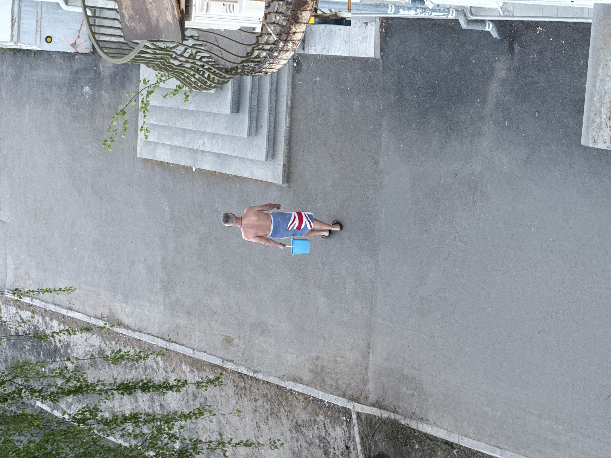 A high-angle shot shows a shirtless man in swim trunks with a Union Jack design, holding a blue object, walking on a paved area. The location appears to be outdoors, possibly a pedestrian area, with steps, metal structures, and trees visible nearby. The exact location in Votkinsk, Russia is not evident from the image. The man is walking away from the steps.