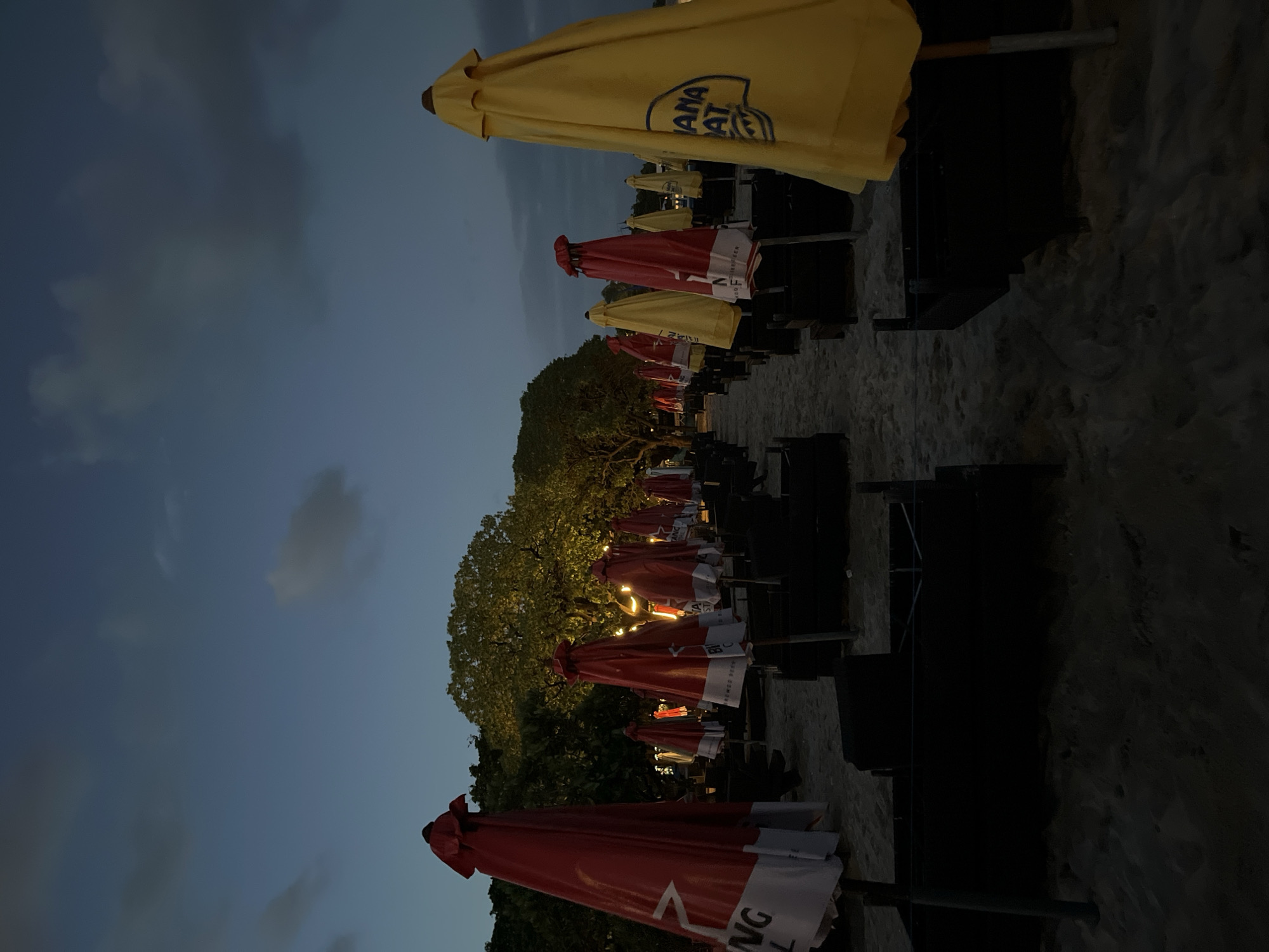 The image shows a row of closed beach umbrellas and lounge chairs on a sandy beach in Kuta Selatan, Indonesia. The umbrellas alternate between yellow with "Banana Boat" branding and red with white lettering. The lounge chairs are dark and appear to be made of metal. The sky is a dusk or twilight blue with some clouds. Trees are visible in the mid-ground, with some string lights illuminating them. The perspective is from a low angle, looking along the row of umbrellas towards the horizon. The beach appears relatively empty.