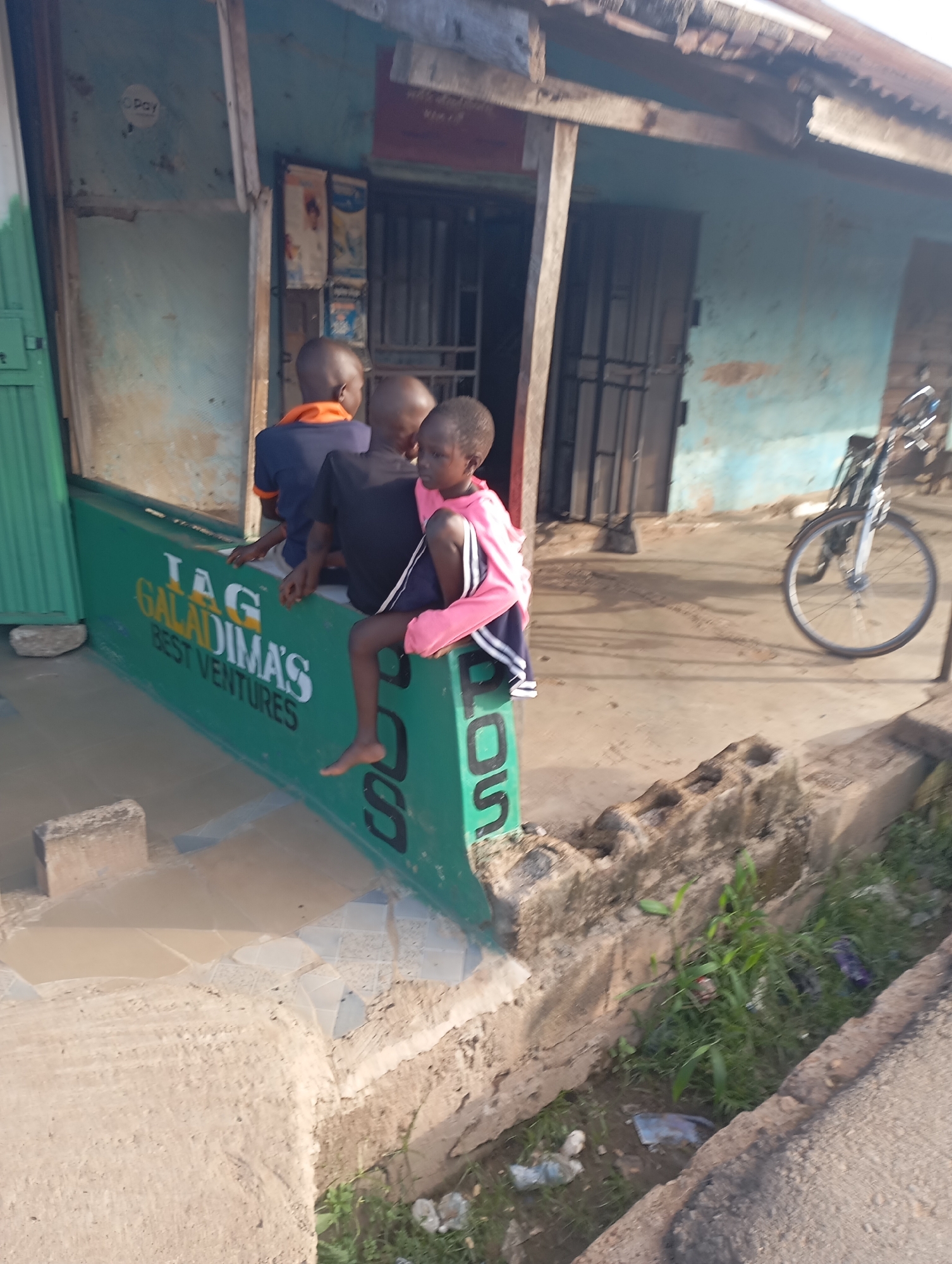 The image shows three young boys sitting on a green sign that reads "IAG GALADIMA'S BEST VENTURES". The boys are of varying ages and appear to be of African descent. One boy in the middle is wearing a black t-shirt and dark shorts. The boy on the right is wearing a pink jacket and dark shorts with white stripes. The boy on the left is wearing an orange and blue t-shirt. They are sitting in front of a building with a light blue exterior. The building has barred windows and a corrugated metal roof. There is a bicycle parked in front of the building to the right. The ground in front of the building is paved with concrete and there is some debris and vegetation. The image appears to have been taken during the day, with sunlight casting shadows on the ground. The location is identified as Shendam, Nigeria.