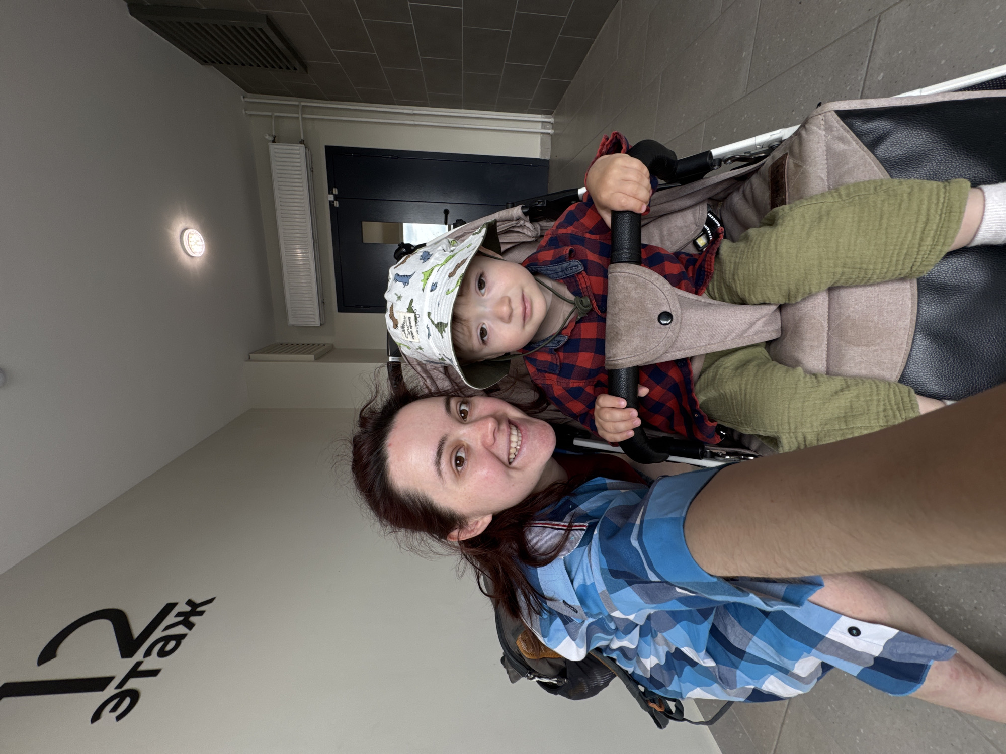 A woman and a young child are in a hallway. The woman is on the left, wearing a blue and white plaid shirt and a backpack. She is smiling at the camera, taking a selfie. The child is in a stroller on the right, wearing a red and black plaid shirt and a bucket hat with dinosaur prints. The child is looking at the camera with a neutral expression. The hallway has a tiled floor and light-colored walls. There is a sign on the left wall that says "12 этаж" (12th floor). In the background, there are double doors, a radiator, and ventilation grates. The overall scene suggests a residential building hallway.