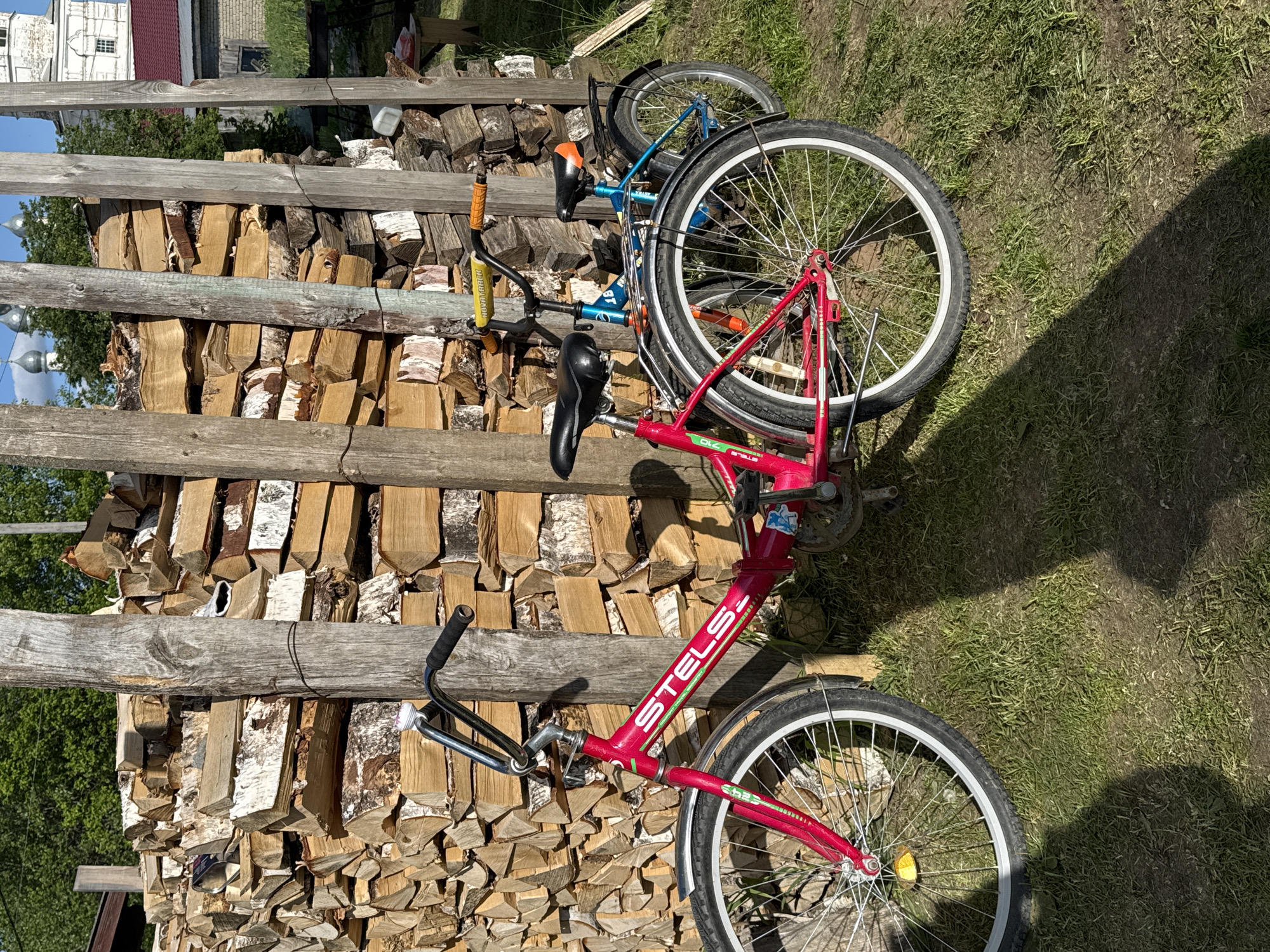 The image, when viewed in its correct upright orientation, captures a scene in a rural or semi-rural setting. Two bicycles are parked side-by-side on green grass next to a large, neatly stacked pile of firewood.
In the foreground, a red bicycle is prominent, featuring a black seat, chrome handlebars, and a white front mudguard. The brand name "STELS" is clearly visible in white letters with green outlines and stripes on its frame. A yellow reflector is attached to the spokes of its front wheel. Behind it, a blue bicycle is partially visible, distinguished by an orange seat and the brand name "NOVATRACK" on its handlebar stem. Both bicycles appear to be suitable for children or small adults.
To the left of the bicycles, a substantial woodpile is stacked high, predominantly composed of split logs, many displaying the characteristic white and black bark of birch trees. The woodpile is situated behind a rustic fence made of rough, horizontal wooden timbers.
The ground consists of green grass interspersed with patches of bare earth. Strong, dark shadows fall across the grass, indicating bright, direct sunlight, suggesting it is midday or early afternoon. In the distant background, partially obscured by trees and the woodpile, are glimpses of buildings, one featuring a white, ornate architectural element that resembles a dome or cupola, visible against a clear blue sky. No people are present in the image.