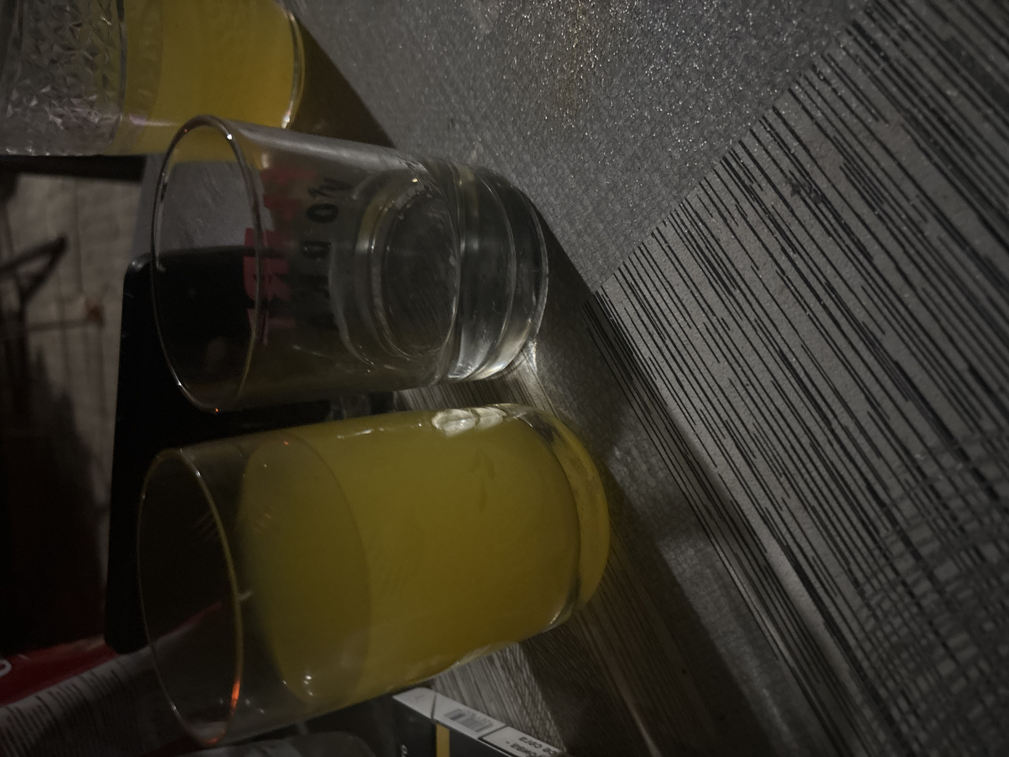 Two glasses are on a table. One glass, on the left, is filled with a yellow liquid. The second glass, to the right, is empty. A pack of cigarettes is partially visible in the lower left corner of the image. The background is dark and out of focus, suggesting an indoor or nighttime setting. The location in Trudovets, Bulgaria, is not discernible from the image.