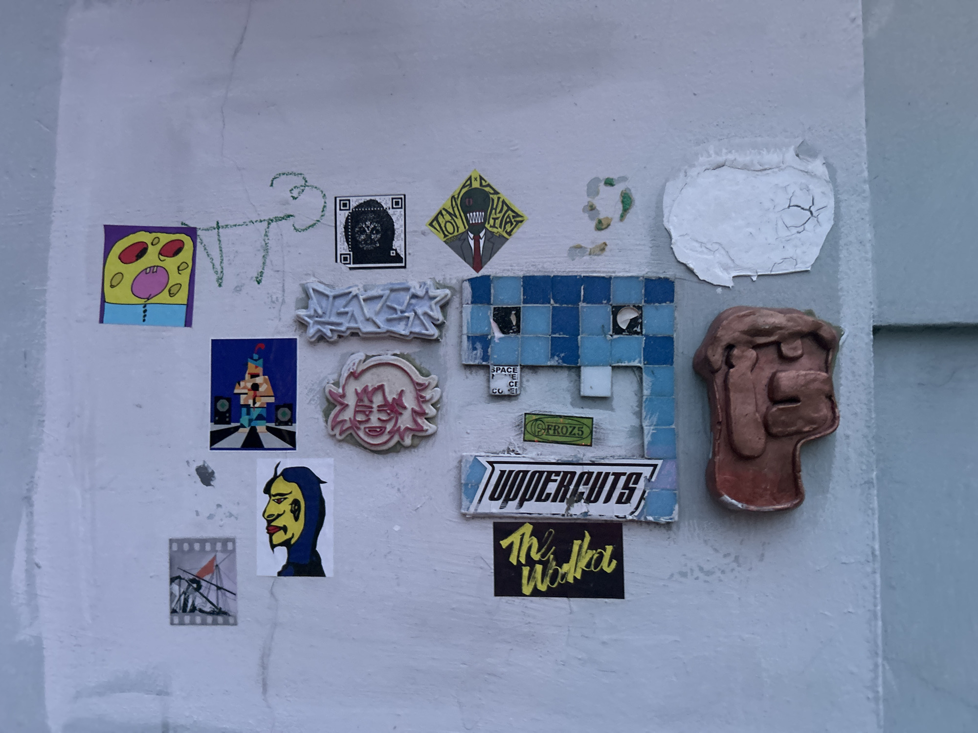 The image shows a section of a light grey wall, likely exterior, covered in various stickers, drawings, and small sculptural elements.
There is a drawing in green marker, and several stickers including: a crudely drawn yellow cartoon figure on purple background; a black and white QR code; a cartoon figure in a suit with a green head and red tie; a tower-like figure on a stage; a yellow-faced figure with a blue hood; a strip of film; a pink-haired cartoon face; text reading "SPACE FACE COFFEE"; "FROZS"; "UPPERCUTS"; and "The Wolka."
There are also three sculptural elements: a white cracked patch, a blue tiled piece, and a dark reddish-brown piece resembling a hand or face.
The location and specific city is unknown.