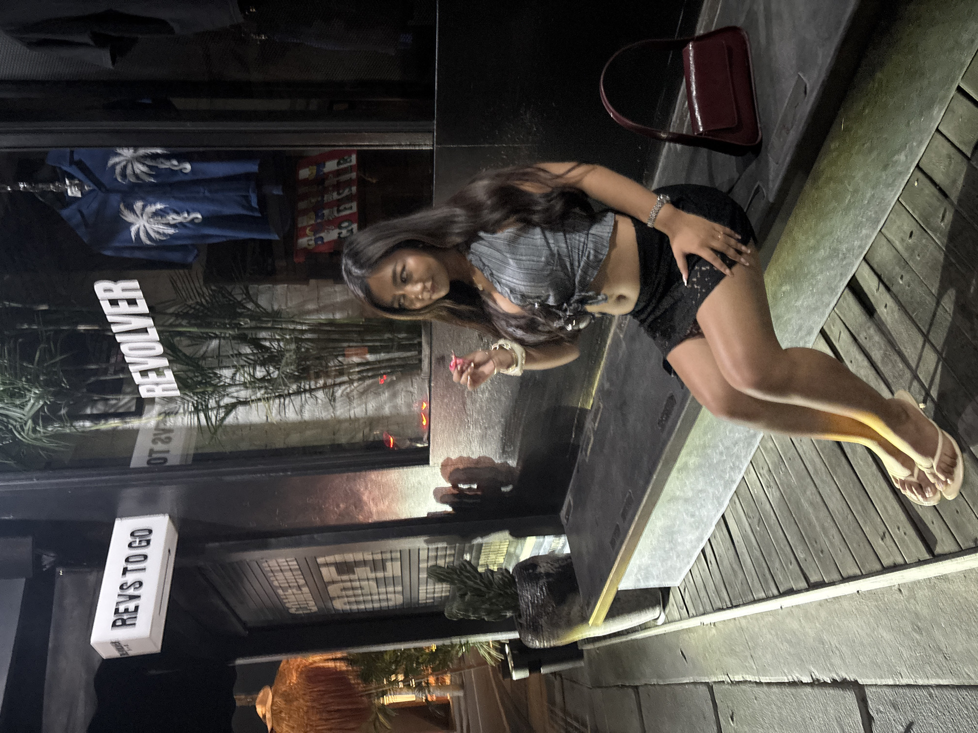 A young woman with long, dark hair is posing for a photo, sitting on a wooden deck. She is wearing a cropped grey and white striped shirt tied at the front and a black skirt. She is holding a pink object, possibly a sparkler or a cigarette, in her right hand, which is adorned with bracelets and rings. Her left hand rests on her thigh.
The setting appears to be outdoors at night, with artificial lighting illuminating the scene. Behind the woman, through a large window, a display of blue shirts with white palm tree graphics is visible. A red purse is placed on the wooden deck to her right. Signs are visible in the reflection of the window; one reads "REVOLVER" and another, a white box, says "REVS TO GO". The wooden planks of the deck are worn and weathered. The overall atmosphere suggests a casual evening setting, possibly outside a shop or restaurant in North Kuta, Indonesia. The woman's expression is relaxed and she appears to be posing confidently.