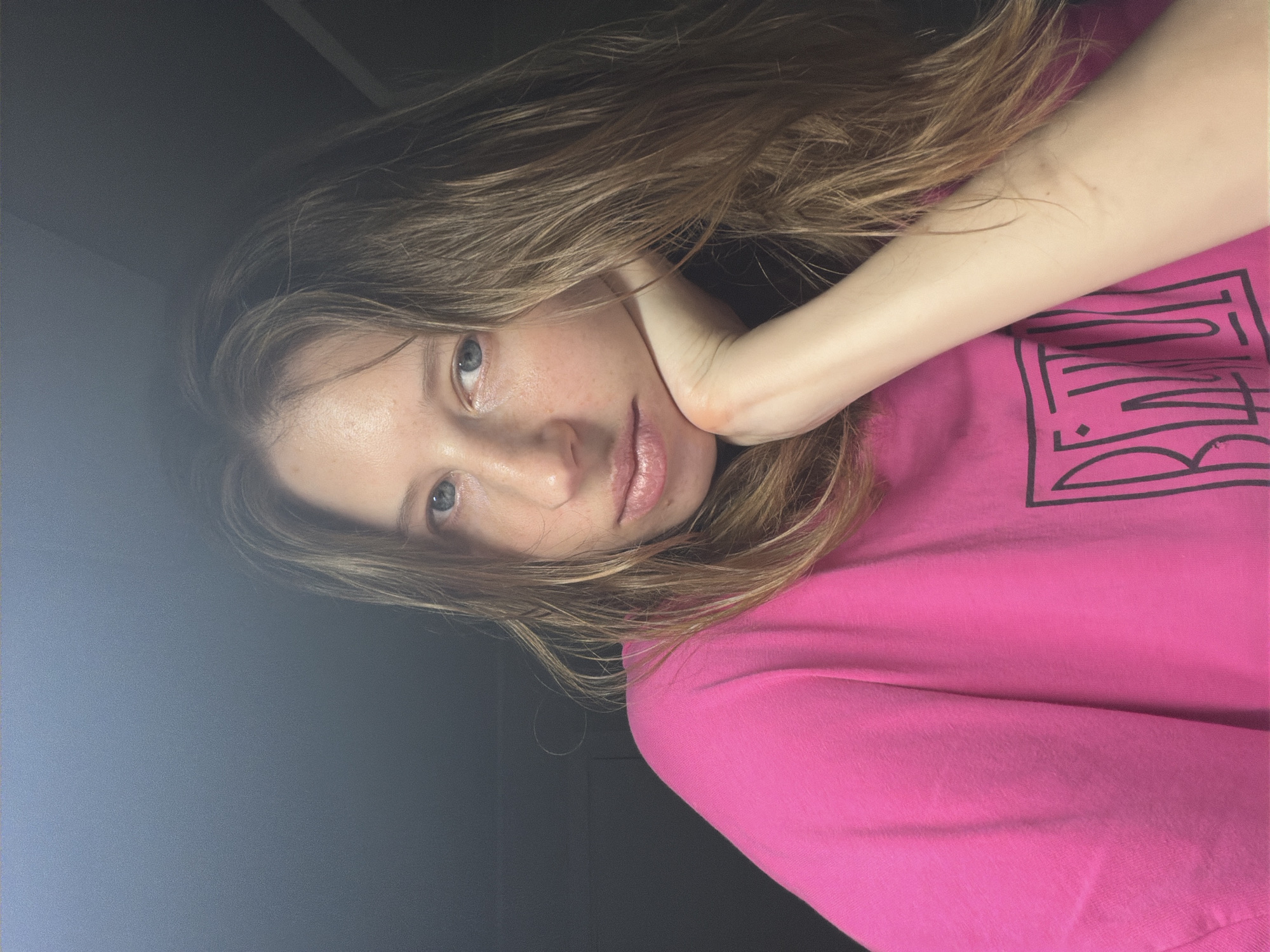 A young woman with light brown hair is captured in a medium close-up shot, looking directly at the camera. She has blue eyes and fair skin, with her right hand resting on her chin, supporting her head. Her hair is parted on the left and falls loosely around her face and shoulders. She is wearing a vibrant pink t-shirt with a black graphic design on the front. The background is dark and indistinct, with a slight gradient from left to right, suggesting an indoor setting with artificial lighting. The lighting highlights her face and upper body, casting a shadow on the left side of her face and neck. The image appears to be taken in Dongducheon-si, South Korea, though no specific city landmarks are visible.