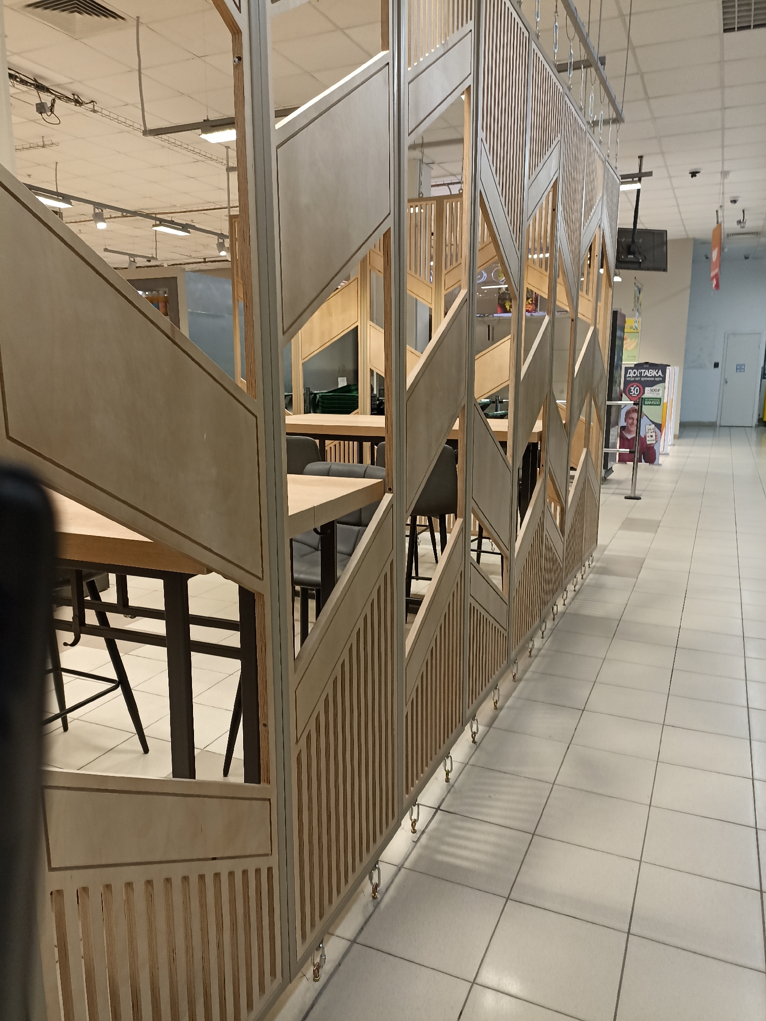 The image shows a modern, indoor cafe or seating area within a commercial space, likely a supermarket or mall, in Moscow, Russia. The area is partitioned by a decorative wooden screen with geometric patterns and vertical slats. Several tables with light-colored wooden tops and dark metal legs are visible, accompanied by dark chairs. In the background, a white tiled floor extends into the distance, with fluorescent lighting fixtures and ceiling panels overhead. Signs in Russian are visible, including one that reads "ДОСТАВКА" (delivery). The scene is clean and well-lit.