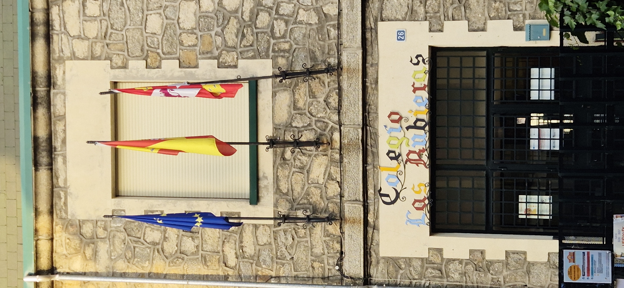 The image shows the facade of a building, likely a school, made of stone blocks. Three flags are mounted on poles outside a window: the European Union flag on the left, the Spanish flag in the center, and another flag on the right with a red and white design. Below the window, there is an archway with the words "Colegio Las Rubieras" written in colorful, decorative lettering. The entrance is a large black metal gate with a grid pattern, and various posters are visible through the glass panels of the gate. A small blue mailbox is mounted on the right side of the wall. There are no people visible in the image.