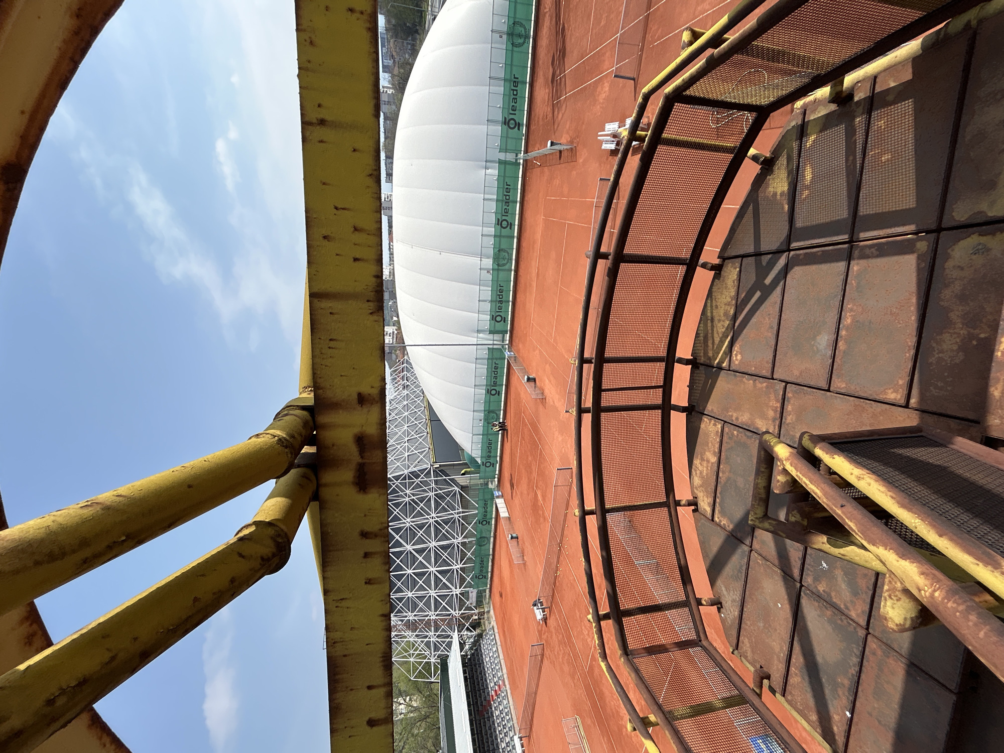 The image shows an elevated view of a tennis court complex in Plovdiv, Bulgaria. A white, domed structure covers one of the courts. The courts are red clay. The photograph appears to be taken from a rusted metal structure, possibly a viewing platform or bridge, with yellow painted supports and metal mesh railings. The sky is blue with some clouds. People can be seen on the tennis courts.