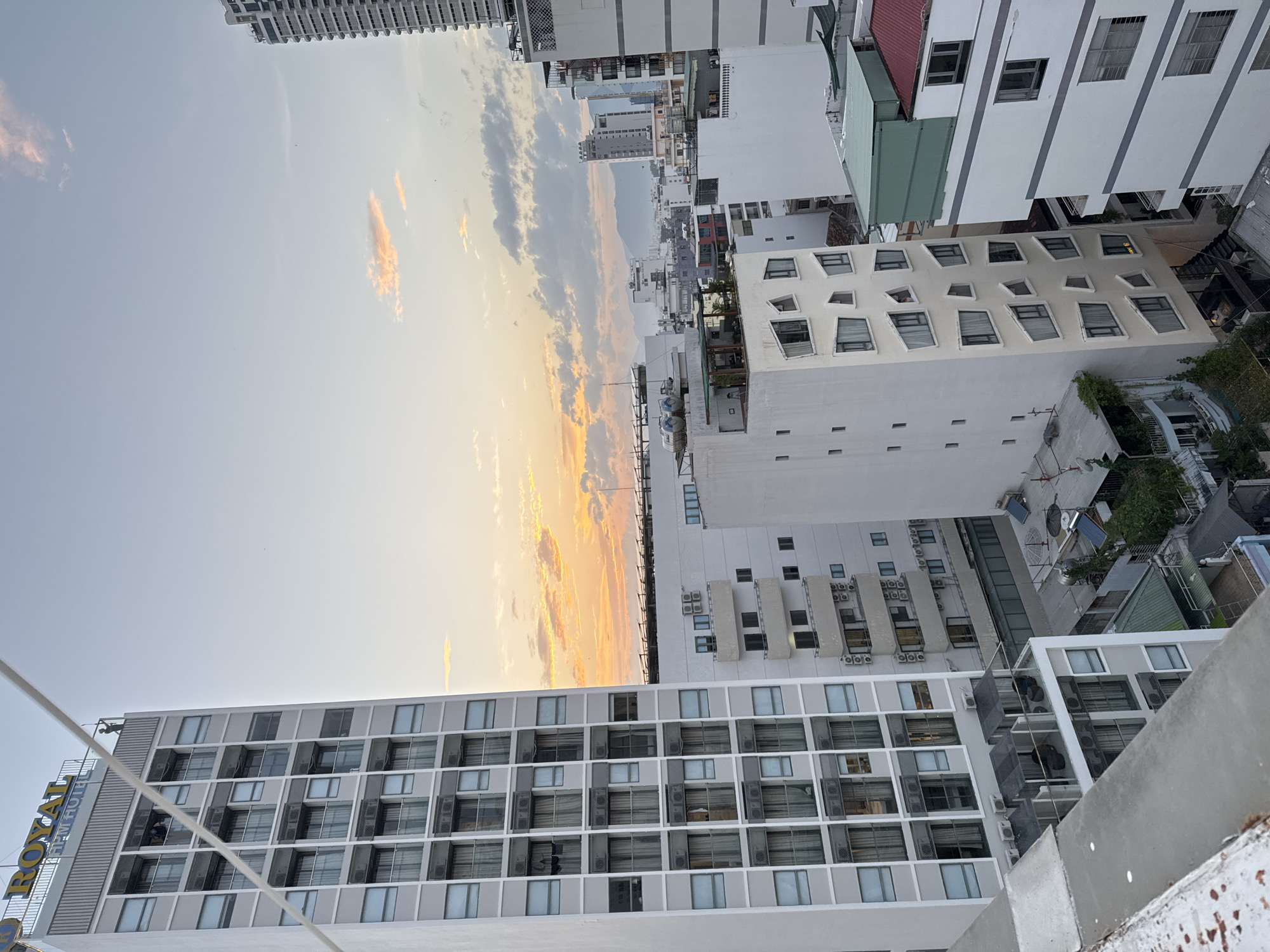 The image shows a cityscape in Nha Trang, Vietnam, with several buildings of varying heights. The sky is a pale blue with scattered clouds, tinged with orange and yellow from what appears to be either sunrise or sunset. In the foreground, a tall, white, multi-story hotel with many windows is visible. A prominent sign at the top of this building reads "ROYAL HPM HOTEL". There are people visible on the rooftop of this hotel, appearing to stand near the edge. To the right and behind the main hotel, other buildings of different architectural styles are present, some with balconies and air conditioning units. The overall scene depicts an urban environment with a view of the sky and distant mountains.