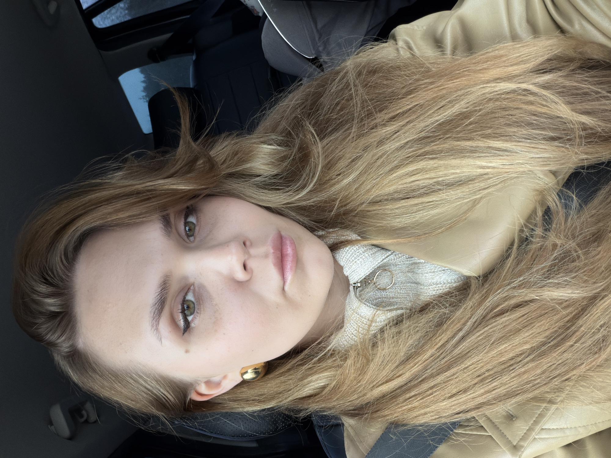 A woman with long blonde hair is in a car. She is wearing a tan leather jacket and a light-colored sweater with a zipper. She has light skin and green eyes. She is wearing a gold earring in her left ear. The car's interior is black. The location is unknown and cannot be identified as being in the city of Прикамский, Russia.