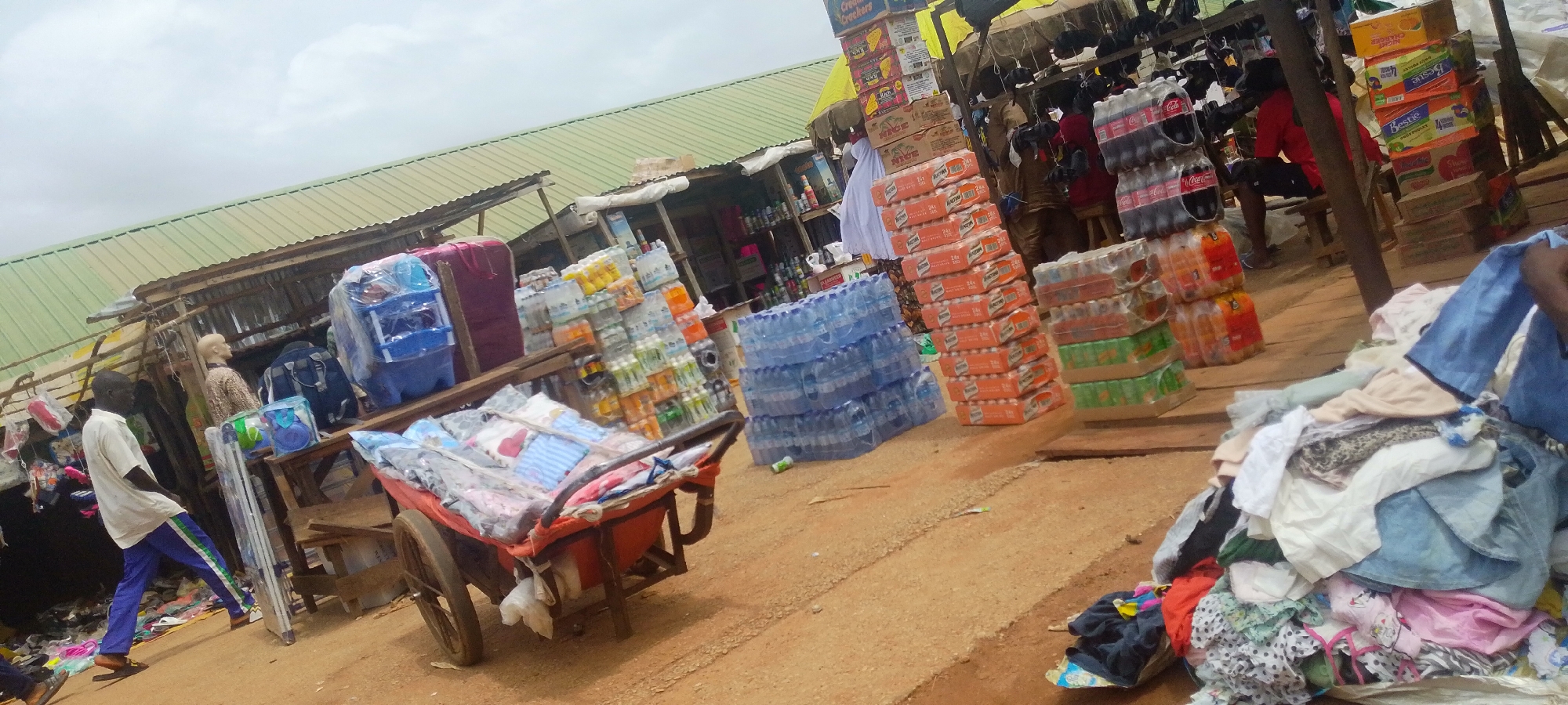 The image depicts an outdoor market scene in Baissa, Nigeria. In the foreground, a man in a white shirt and blue pants pushes a red cart loaded with folded fabrics and clothing. Behind him, a market stall displays various goods, including stacks of bottled water, cases of soda, and boxes of packaged food. A blue rack holding more items is also visible. Further in the background, the market extends with more stalls covered by green corrugated metal roofs. People are present, including a man in a red shirt sitting on a stool, and another person, possibly a vendor, partially visible behind the stacks of drinks. The weather appears overcast, suggesting it might be daytime with cloudy skies. The setting is characteristic of a bustling local marketplace, with goods piled high and people engaged in commercial activities. There are several instances of visible text on product packaging, such as "Coca-Cola" on the soda cases and labels on the food boxes, though most of the text is not legible. The overall impression is one of everyday commerce in a Nigerian town.