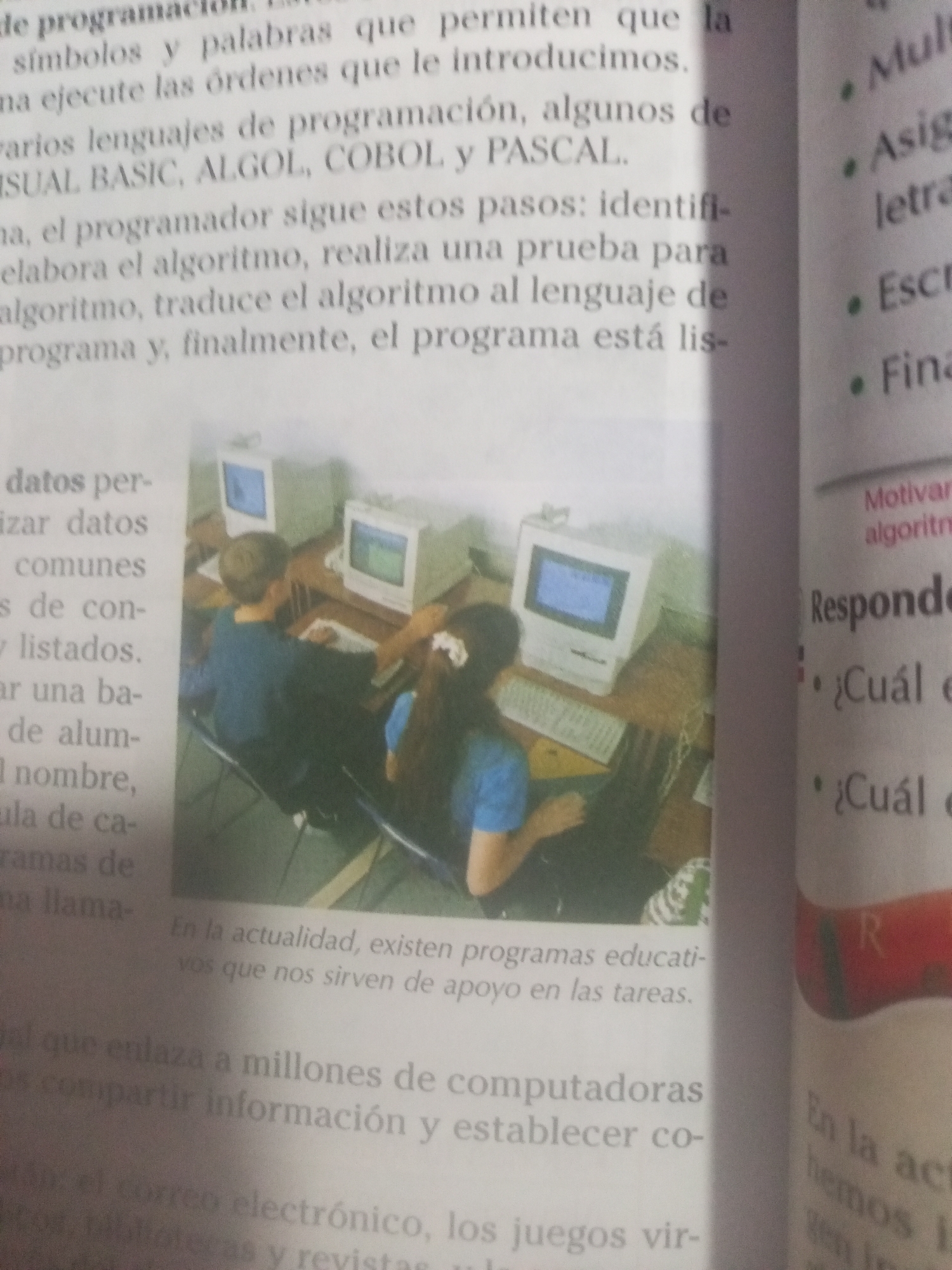 The image shows a page from a book or magazine with text and a photograph. The text discusses programming languages and the steps involved in creating a program. The photograph depicts two children, a boy and a girl, sitting at computers in a classroom or computer lab. The boy is wearing a blue shirt and the girl is wearing a blue t-shirt. Both are looking at their computer screens and appear to be engaged in a learning activity. The background shows more computer equipment and desks. The image is in color and appears to be well-lit. There is no information in the image that indicates a specific location in La Tagua, Venezuela.
