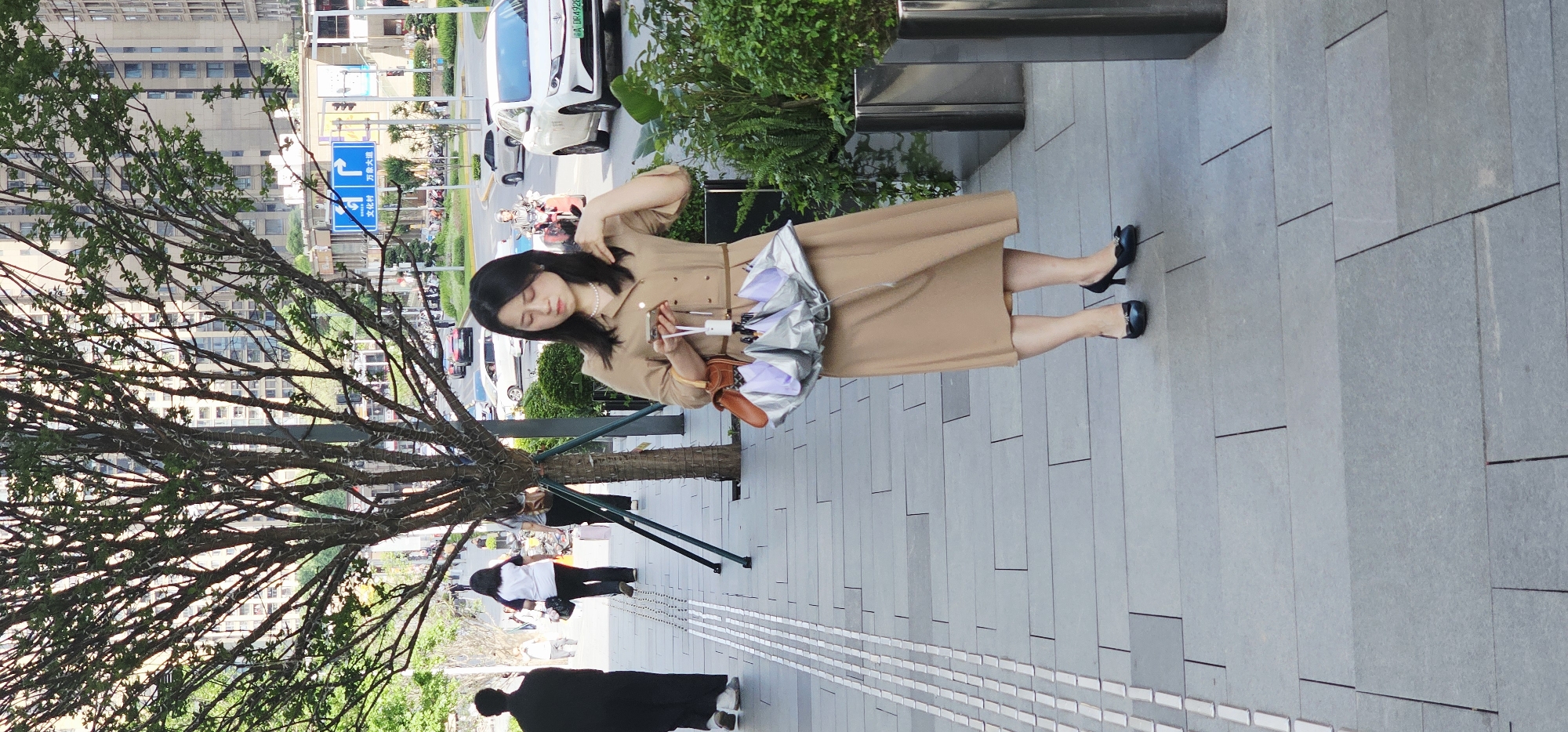 A young woman is depicted standing on a paved sidewalk in a daytime urban setting, likely in Jiulongpo District, China. She is positioned towards the right side of the frame, looking down at a smartphone held in her hands. She wears a knee-length, light brown or tan shirt dress, black high heels with decorative bows, and a pearl necklace. Her dark hair falls around her shoulders, and one hand is near her hair. A distinctive silver umbrella with purple accents is attached to her side, seemingly by a brown, leather-like pouch or strap.
To the left, a tree with numerous intricate, bare branches partially obscures the background. Beyond the sidewalk, a busy street is visible with several vehicles, including white cars and motorcycles, and tall multi-story buildings further in the distance. Lush green foliage and planted areas are situated between the sidewalk and the street.
Other people are present in the scene, including a figure dressed in black in the lower left foreground and another person in a white t-shirt and dark pants standing near the tree in the mid-ground.
Notable details include a blue street sign with white text displaying Chinese characters: "文化村" (Wenhua Village/Culture Village) and "万寿大道" (Wanshou Avenue), accompanied by a left-turn arrow. A car's license plate in the background is partially visible, starting with "渝A J9R...", indicating a Chongqing registration. The lighting suggests a bright day, possibly sunny or lightly overcast.