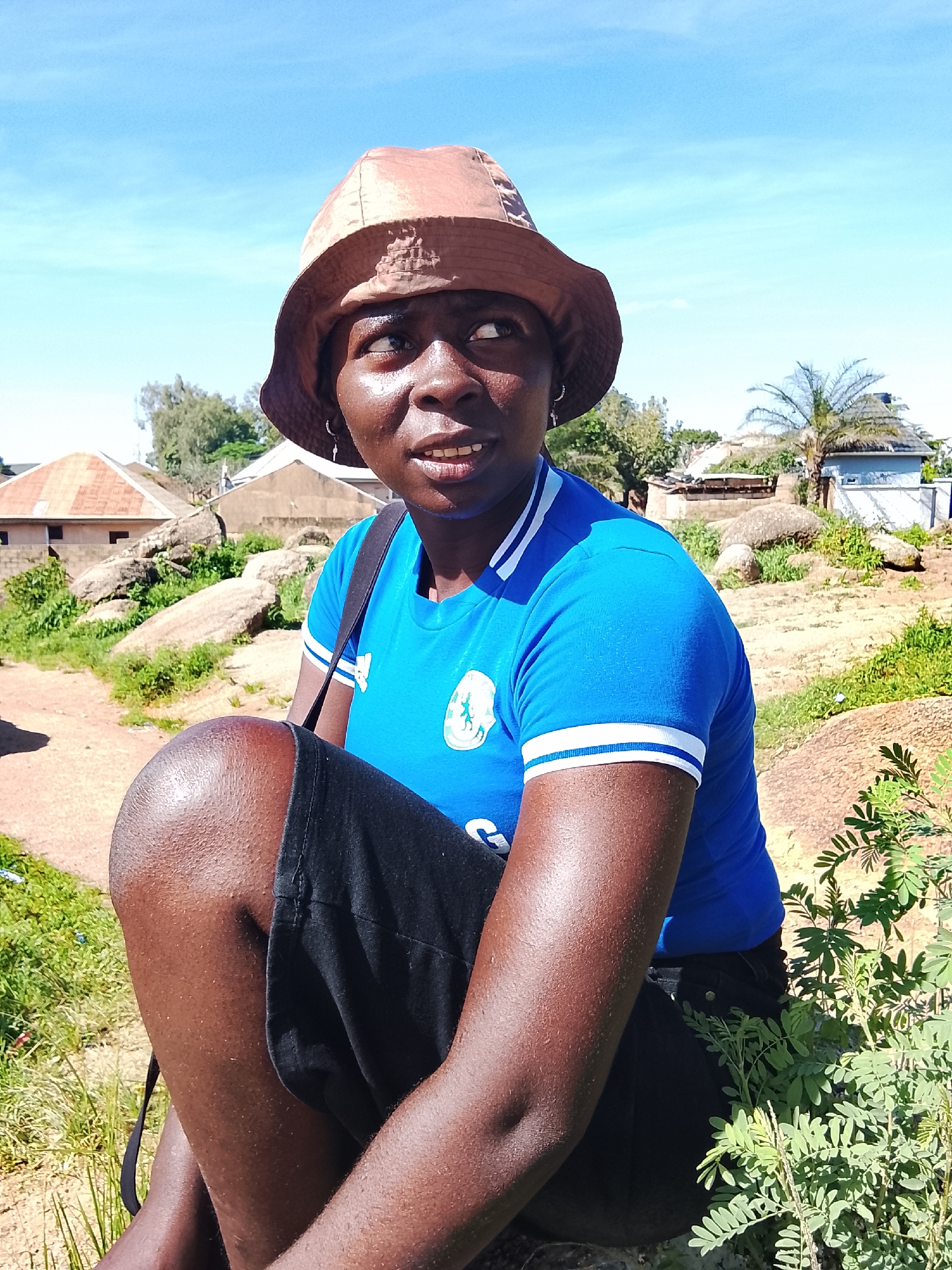 A medium shot captures a Black woman wearing a brown bucket hat and a bright blue t-shirt with white stripes on the sleeves, and black shorts. She is sitting on a large rock, with her left knee bent and pulled up towards her chest. The woman is looking up and to her right with a slight smile. Her skin is a deep brown, and her face is glistening with sweat.
The background is a landscape with rocky terrain and sparse vegetation, under a clear blue sky. Several small buildings with corrugated metal roofs are visible in the middle ground, suggesting a semi-urban or rural setting. The buildings appear to be simple, with muted earth tones. The ground is a reddish-brown dirt path. A small green bush with delicate leaves is in the foreground on the right side of the frame.
The lighting is bright and appears to be natural sunlight, creating distinct shadows and highlights on the woman's face and arms. The overall impression is that of a warm, sunny day, likely in an outdoor environment in Jos, Nigeria. The woman's attire and the setting suggest a casual and possibly active moment, perhaps during a break or while exploring the area.