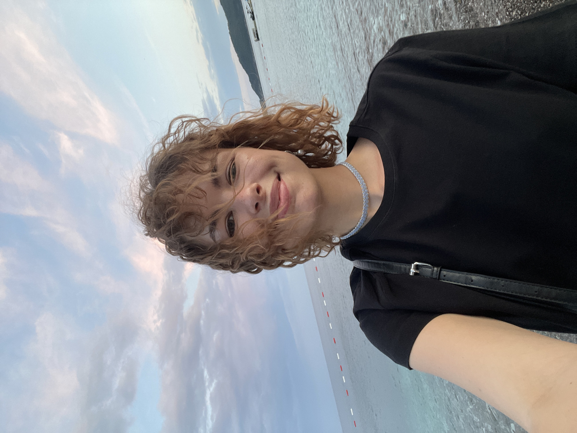 A rotated outdoor selfie photograph features a woman in the foreground, wearing a black T-shirt and a light blue choker necklace. A black shoulder bag strap is also visible. She has curly brown hair and a smiling expression.
The background is a coastal scene, likely a beach. The sky is visible above, displaying a mix of pale blue and white clouds, suggesting either dawn or dusk.
The location is identified as Rafailovići, Montenegro. The water appears calm, and there are what seem to be markers in the water in the distance. There is land visible in the background, likely hills or mountains.