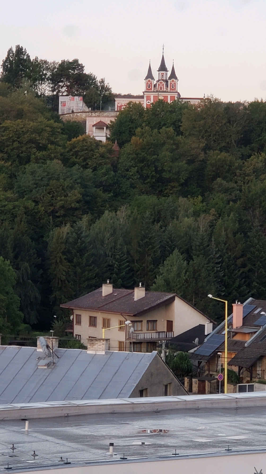 In Prešov, Slovakia, an elevated view captures a residential area nestled against a densely forested hill. At the summit of the hill, a prominent red and white building with multiple spires, resembling a church or monastery, stands against a pale sky. Below this structure, a lighter colored building with a small turret is partially visible, surrounded by scaffolding, suggesting ongoing construction or renovation. The hillside is densely covered with a variety of trees, creating a verdant backdrop. In the foreground, several houses with pitched roofs are visible. One house has a brown roof and light beige walls with multiple windows. Another house, closer to the viewer, has a grey, sloped roof and dark grey walls. Solar panels are visible on the roofs of some structures further back. A bright yellow street lamp stands prominently in the mid-ground. The scene depicts a typical European townscape, blending urban dwellings with natural surroundings.