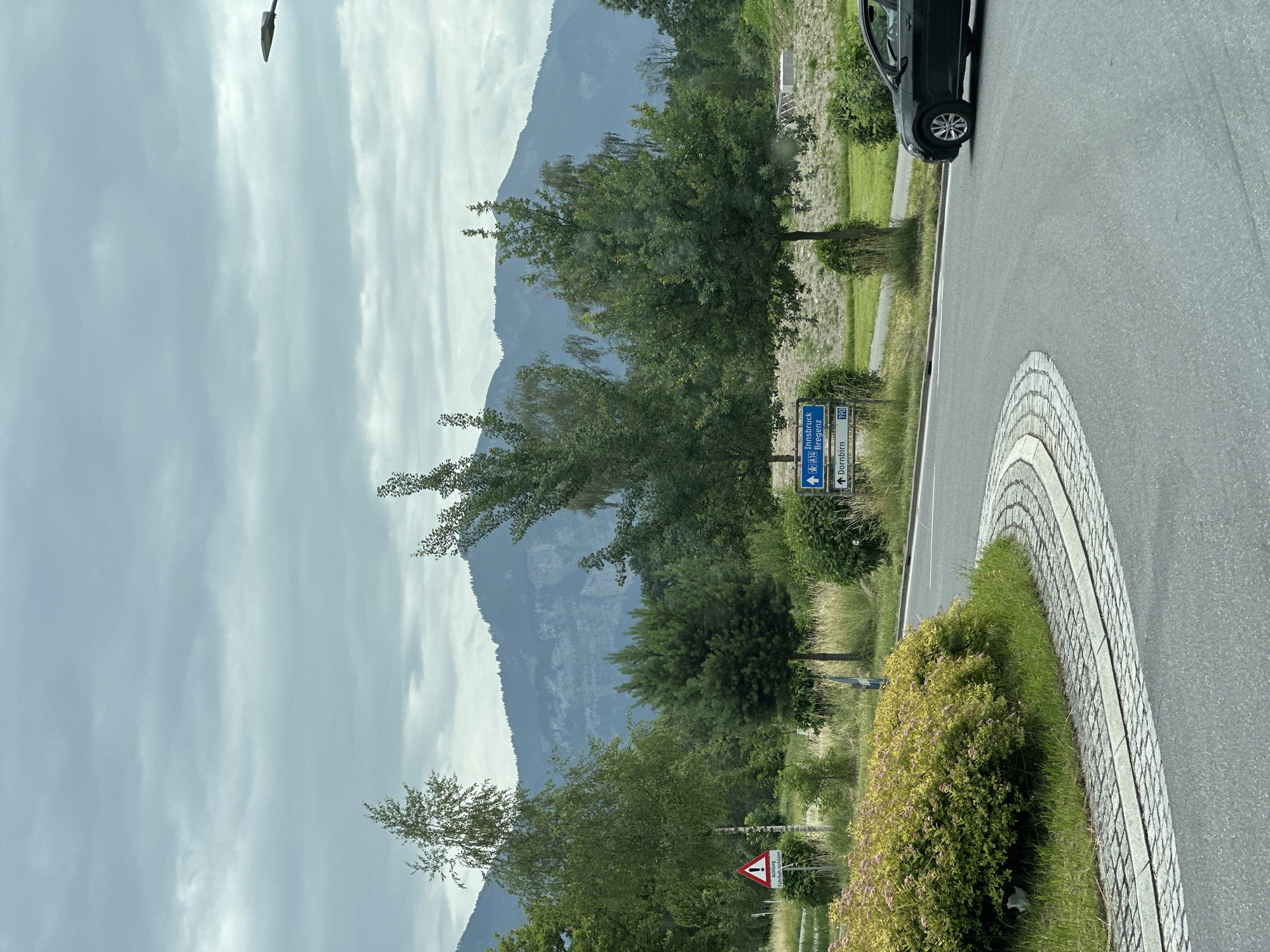 The image depicts a slightly overcast day in Wolfurt, Austria, featuring a road curving to the right with a black car on it. In the foreground, a roundabout with decorative paving stones and a lush green and yellow bush is visible. Beyond the roundabout, the landscape transitions to a field with tall grass, bordered by various trees. In the middle ground, a blue directional sign indicates "Innsbruck A14 Bregenz" and "Dornbirn 190." Further back, a series of hills and mountains form the backdrop under a cloudy sky. A smaller sign with a red exclamation mark and German text is also present on the left side of the image.