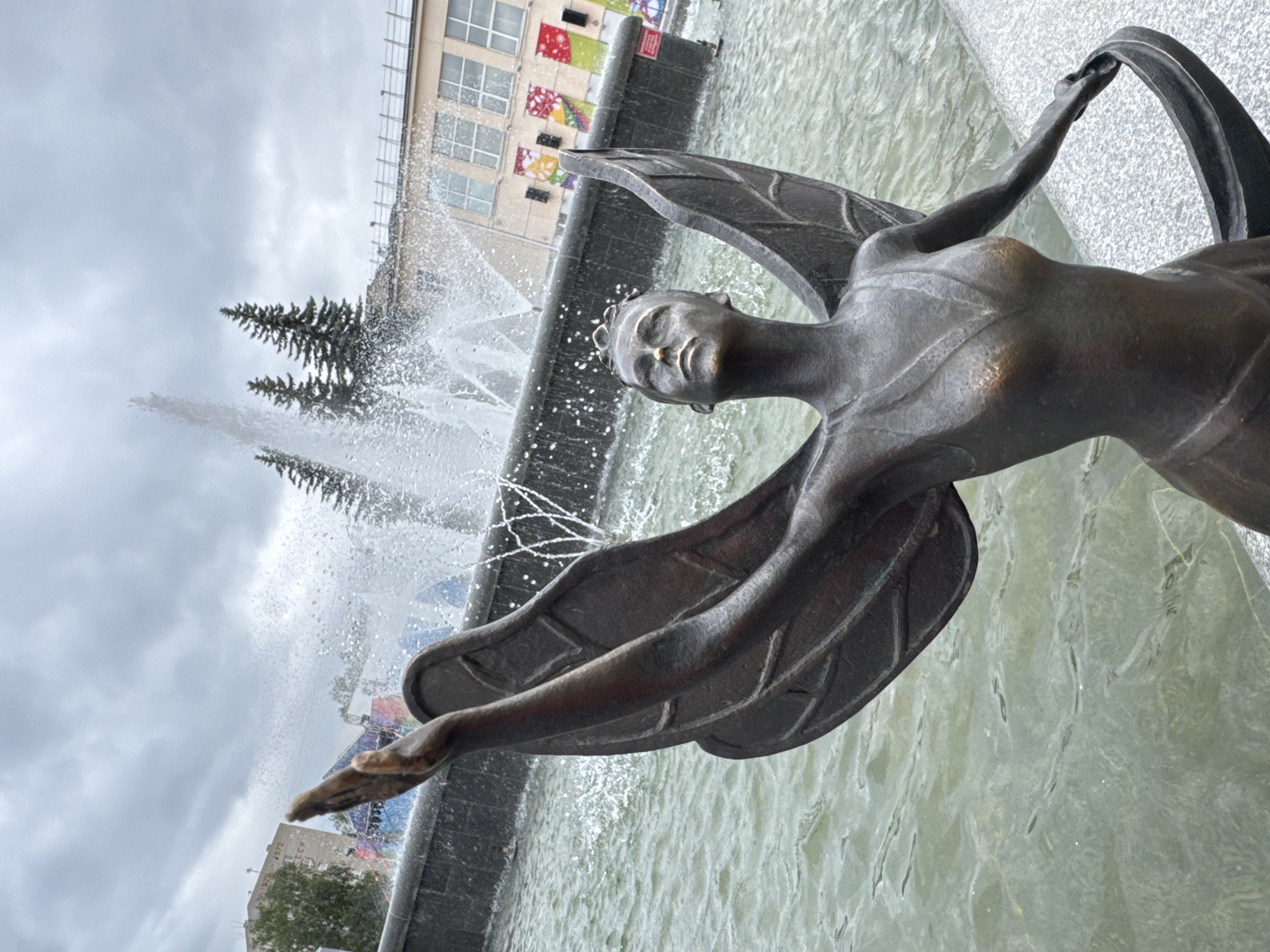 The photograph depicts a bronze statue of a winged woman with her arms outstretched, positioned in front of a fountain. The statue is made of dark bronze and has visible wings extending from its back, as well as intricate detailing on its clothing and face. The woman has her head tilted upwards, looking towards the sky.
Behind the statue, a fountain is actively spraying water, creating a dynamic and misty atmosphere. The water is a greenish hue, suggesting it is a pool or basin. In the background, there are buildings and evergreen trees. The sky is overcast with clouds, indicating a possibly cool or rainy day. The overall setting appears to be an outdoor public space, likely a park or plaza.
This statue is located in the city of Chaikovsky, Russia, specifically in the central city park, known for its fountains and recreational areas. The statue is a prominent feature of the park, often admired for its artistic merit and its symbolic representation.