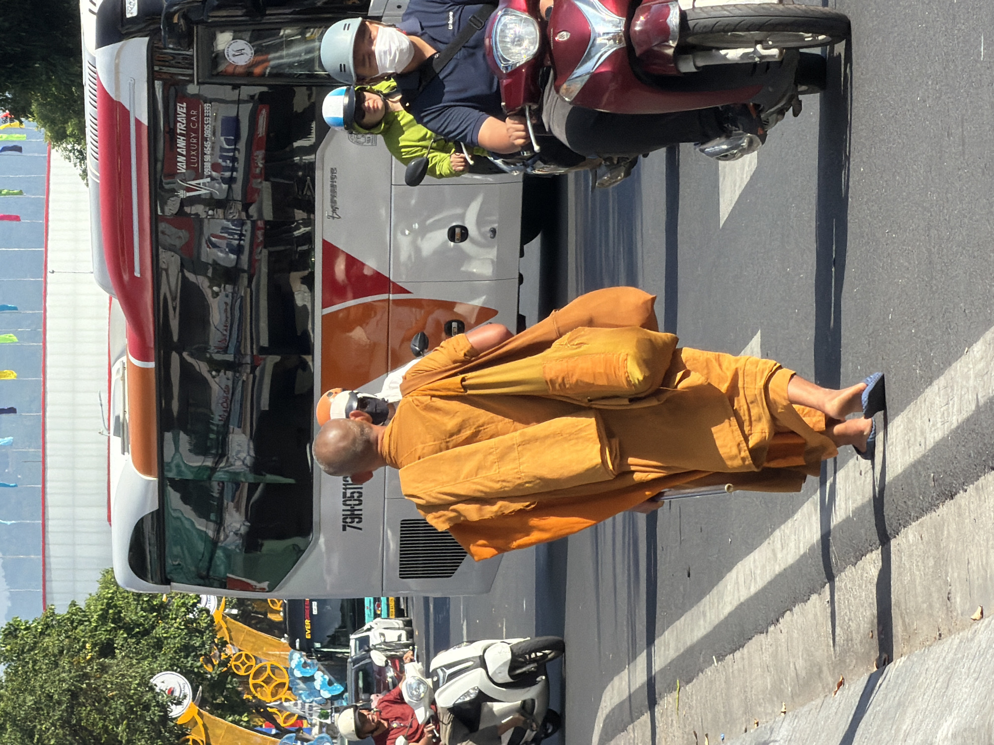 In Nha Trang, Vietnam, on a sunny day, a monk wearing an orange robe walks along the side of a busy street, carrying a yellow bag over his shoulder. He is walking away from the camera, towards a white bus with orange and red accents, which has the text "VAN ANH TRAVEL LUXURY CAR" on its side. Several vehicles are present, including a white scooter to the left with a person wearing a red shirt and white helmet, and a red scooter to the right with two people wearing helmets and one wearing a face mask. The street is paved and marked with white lines. Trees and flags are visible in the background.