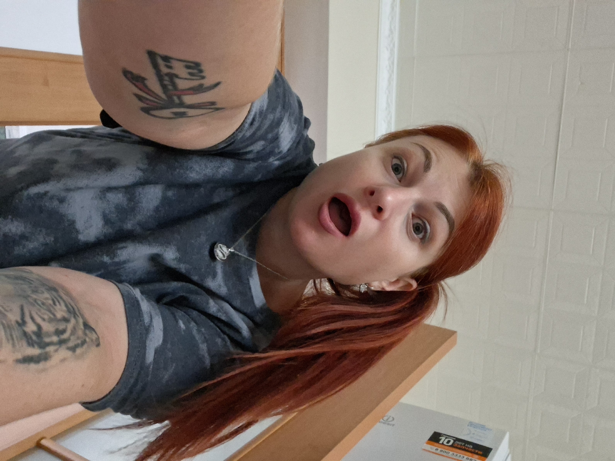The image shows a woman with bright red hair pulled into a ponytail, taking a selfie. She is wearing a dark gray tie-dye t-shirt and a silver necklace with a circular pendant. Her mouth is open in a surprised expression. She has a tattoo on her left arm that appears to be tribal or symbolic, and a tattoo of a tiger's face on her right arm. The background is slightly blurred, but appears to be indoors, possibly a staircase or hallway with light-colored walls and ceiling tiles. A white box with orange lettering is visible in the upper right corner. The image does not provide any information that would allow for localization in the city of Myadzyel, Belarus.