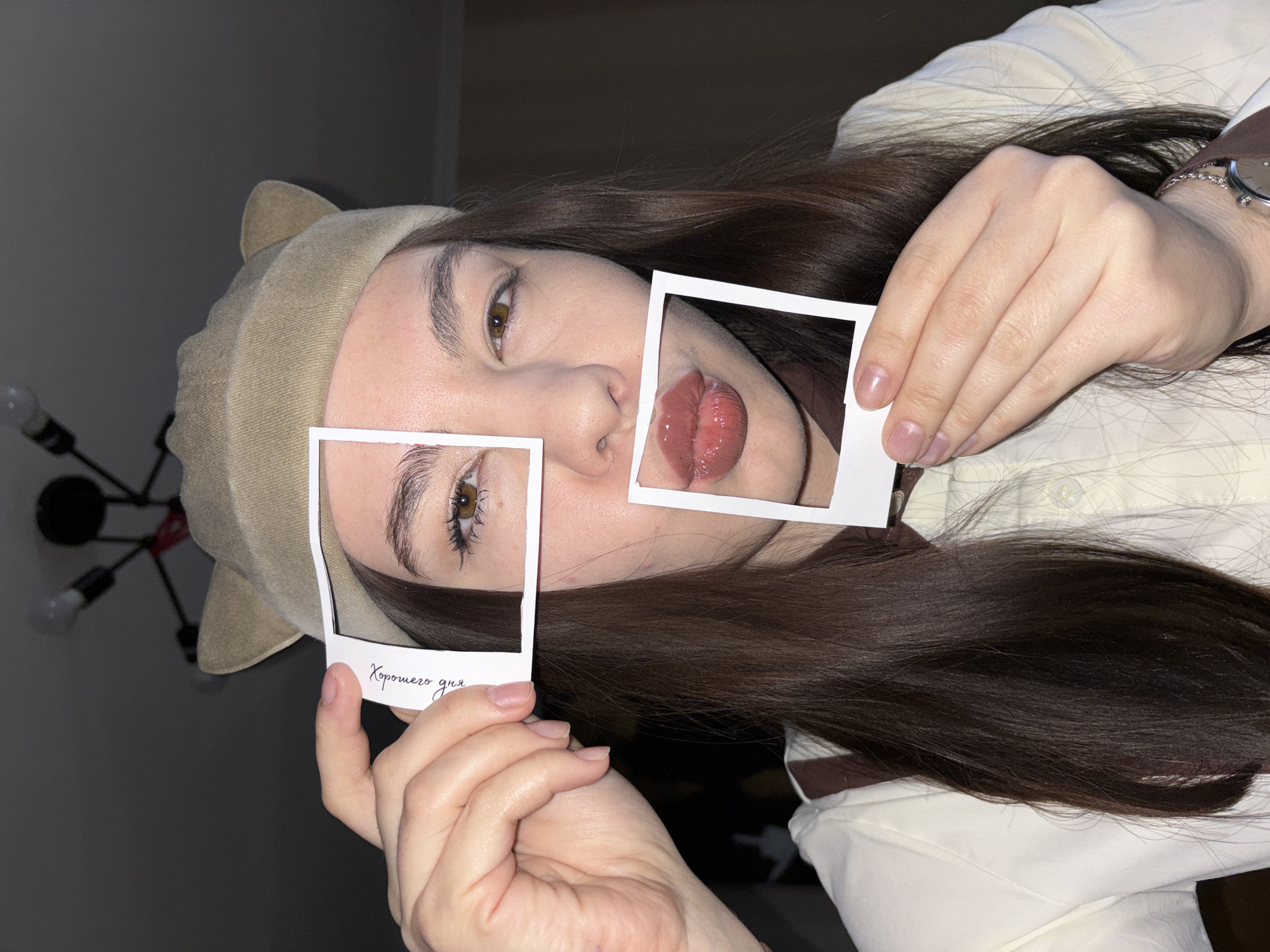 A medium close-up, eye-level portrait shot in Perm, Russia, shows a person with long, dark brown hair, lying down or reclining indoors. The subject wears a light-colored shirt and a light brown hoodie with small, pointed ears on the hood. They are holding two small, white instant photo frames in front of their face. One frame highlights their left eye, which is a warm brown color with visible eyelashes, and the other frame strategically frames their glossy, pinkish-brown lips. Their gaze is directed towards the camera.
The overall lighting is dim, suggesting an indoor setting with artificial light, possibly in the evening. In the blurry background, a distinctive modern chandelier with exposed light bulbs is visible. On the person's right wrist, a silver-toned watch with a dark brown strap is worn. The hands holding the frames have naturally shaped nails.
The lower border of the left photo frame features handwritten text in Cyrillic, "Хорошего дня," which translates to "Have a good day." The image conveys a casual and artistic tone, emphasizing facial features through the creative use of the photo frames.