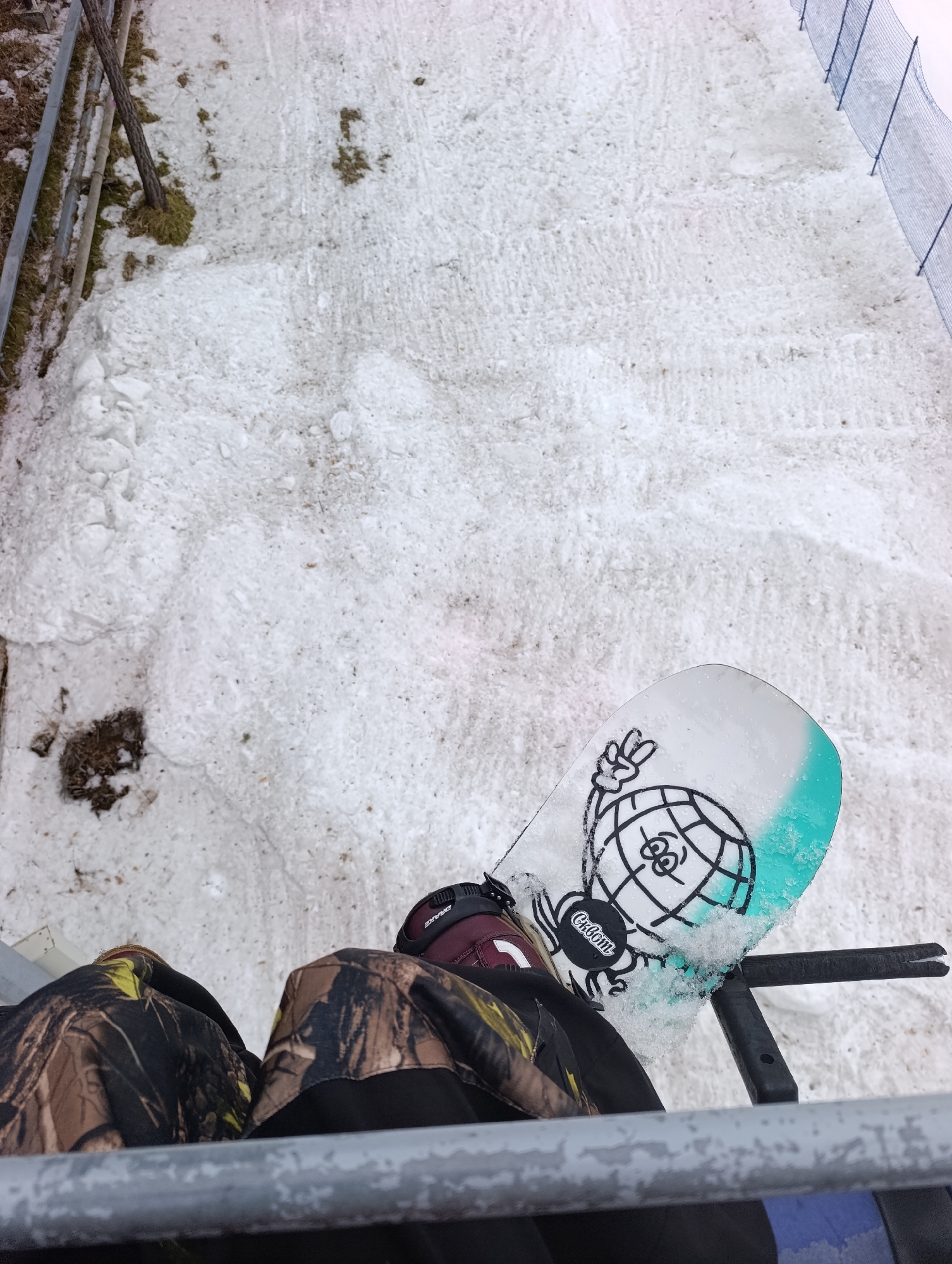 The photograph appears to be taken from a chairlift at a ski resort. The primary subject is a snowboard resting on the chairlift's footrest. The snowboard has a graphic design on its surface, including a stylized globe and a "peace" sign. The tip of the board has a blue/green color. The person in the image is wearing camouflage-patterned pants and ski boots. The background consists of a snow-covered slope. To the right is a blue mesh safety net. The location is likely a ski resort near Krasnoyarsk, Russia, based on the user's city information.
