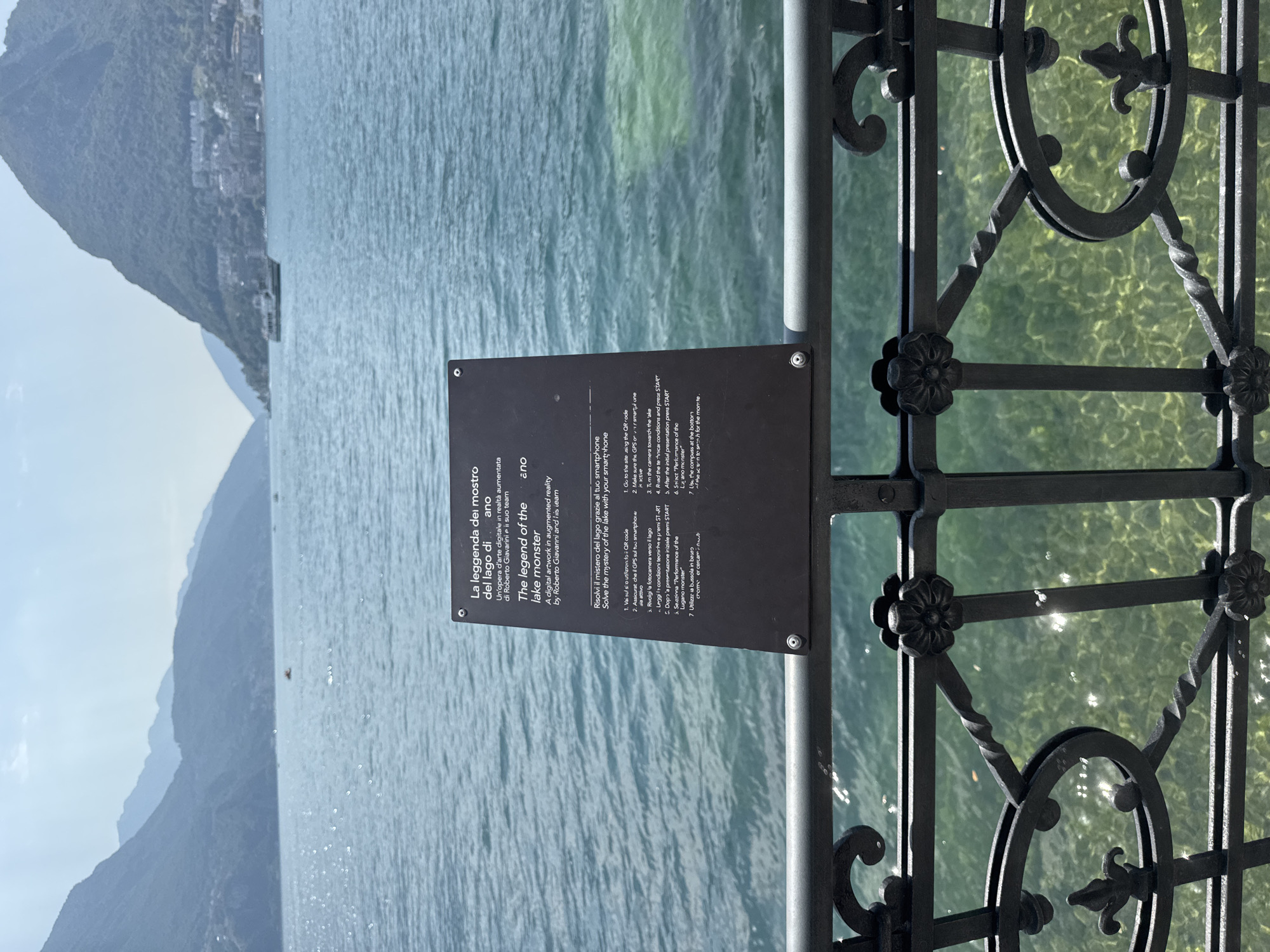 A black metal sign is mounted on a railing overlooking Lake Lugano in Switzerland. The sign is in both Italian and English and describes an augmented reality artwork called "The legend of the lake monster" by Roberto Giavarini and his team. The artwork is described as a digital artwork in augmented reality. The sign provides instructions on how to solve the mystery of the lake with your smartphone. The background features a serene lake with distant mountains and a boat. The railing is made of dark metal with decorative floral and scrollwork elements. The water is clear and turquoise, allowing a view of the lakebed.
