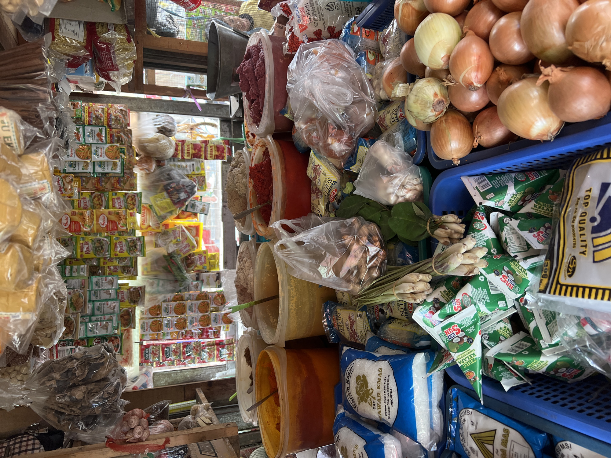 The image shows a market stall in Citeureup, Indonesia, displaying a variety of food items. The stall is filled with shelves and baskets containing packaged goods such as rice, spices, and condiments. Several large plastic containers hold bulk ingredients like chili paste, ground spices, and what appears to be meat paste. Bags of dried goods, possibly mushrooms or herbs, hang from the ceiling. Lemongrass, garlic, and onions are also visible, some are bundled and others in plastic bags. Packaged noodles and rice are stacked in blue plastic crates. The overall scene is one of a busy, well-stocked local market.