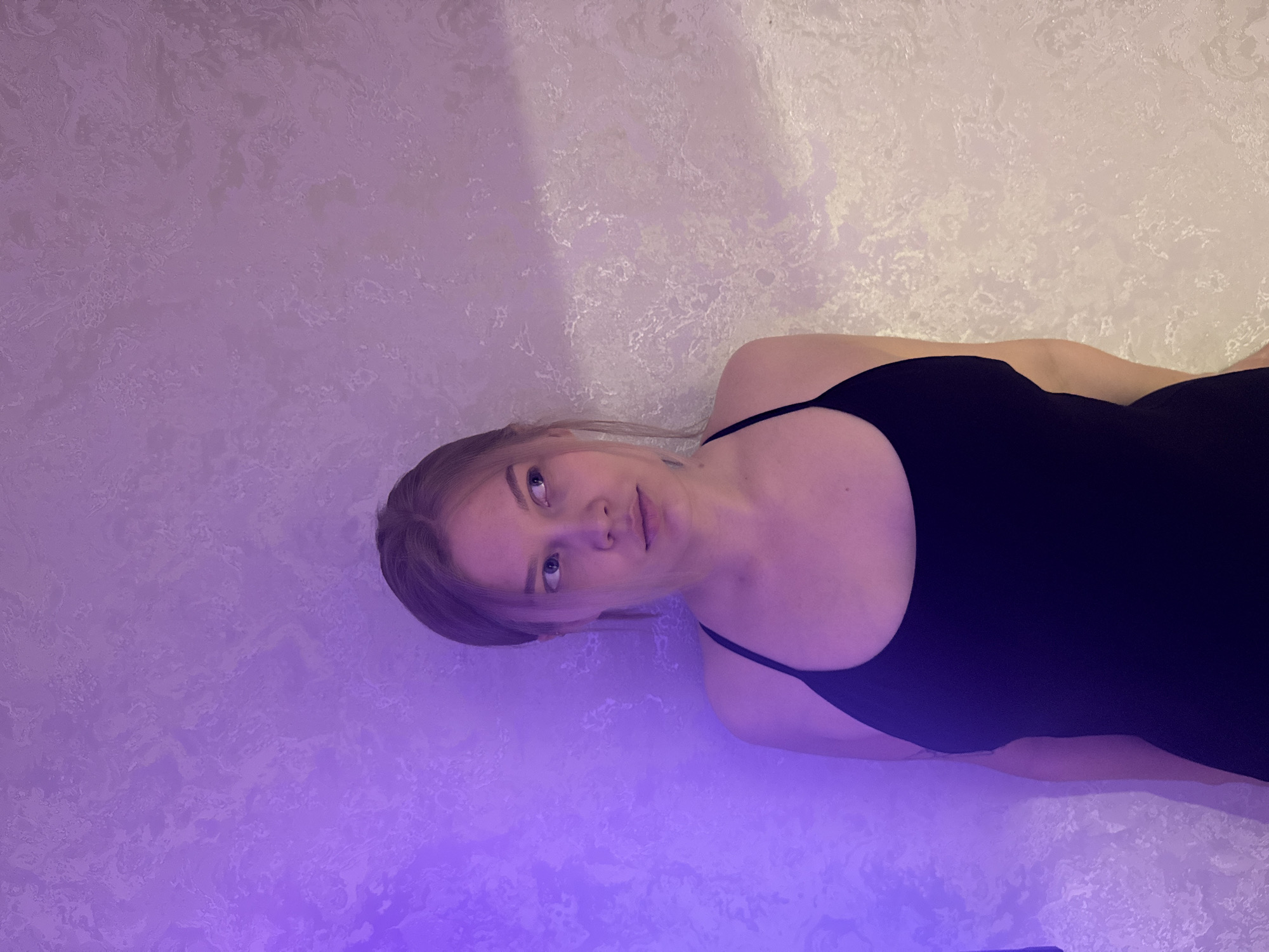 The image captures a portrait of a young woman taken indoors, likely in Chaikovsky, Russia. The woman is positioned as if leaning or lying against a textured wall, but the image is rotated 90 degrees clockwise, so she appears to be looking upwards from the right side of the frame. She has light-colored hair, possibly blonde or light brown, partially pulled back, with some strands falling across her forehead. Her eyes are light, and she looks directly towards the camera with a neutral to slightly contemplative expression.
She is wearing a simple dark-colored, likely black, sleeveless top or swimsuit with thin straps. Her shoulders and upper torso are visible.
The background is a wall with a mottled, textured finish, appearing in varying shades of light purple and white. The entire scene is bathed in a strong, artificial purple or lavender light, which casts a vibrant hue over the wall and her skin, creating a distinct, somewhat atmospheric mood. There is a brighter, almost white area on the right side of the wall, and the purple light seems more intense towards the bottom left. The overall lighting suggests an indoor setting, possibly during the evening or night, using colored mood lighting. There are no identifiable objects besides the woman and the wall. No text is visible in the image.